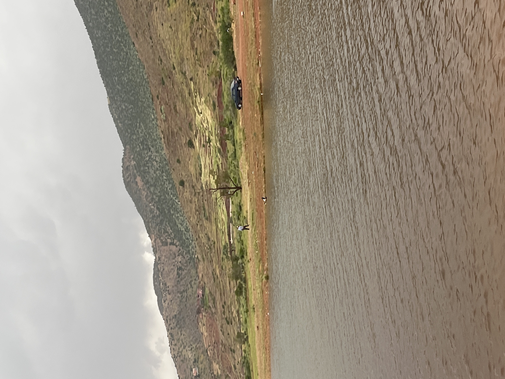 This image, taken in Bin El Ouidane, Morocco, captures a panoramic view of a reservoir or lake bordered by a mountainous landscape under an overcast sky.
The right half of the frame is filled by the expansive body of water, characterized by its brownish-grey hue and subtle ripples on the surface. A narrow, reddish-brown dirt path or bank runs alongside the water. Two individuals are visible on this bank: one person wearing a white top stands closer to the foreground, while another smaller figure is further down the path. A dark-colored car, appearing to be a sedan or SUV, is parked directly on this dirt path near the water's edge. A section of a bare tree trunk or branch is noticeable in the lower-left portion of the frame.
Beyond the shoreline, the terrain ascends into rolling hills and mountains. The immediate slopes show a mix of reddish-brown earth and sparse green vegetation. Further into the background, the mountains become more prominent and are densely covered with dark green foliage. The sky above is entirely overcast, appearing grey and cloudy, suggesting a muted daylight setting. The scene conveys a sense of natural tranquility, with human presence marked by the parked car and the two individuals on the bank. There is no visible text in the image.
