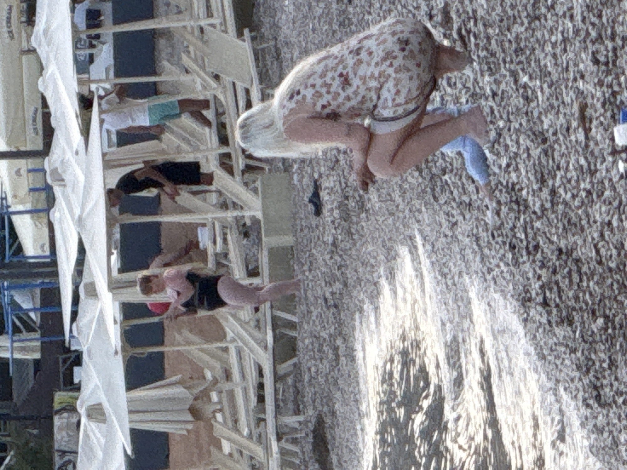 In Budva, Montenegro, on a pebble beach, two women are visible. The woman in the foreground, facing away from the camera, has blonde hair and is wearing a light-colored sleeveless dress with a floral pattern and light blue bottoms, possibly shorts or leggings. She is crouched down. Behind her, a woman with short brown hair, wearing a black one-piece swimsuit and a pink top over it, is standing and holding a red ball. Several rows of white lounge chairs and umbrellas are present on the beach, with some people standing and walking among them in the background. The sea is visible on the left side of the image, with sunlight reflecting on the water.