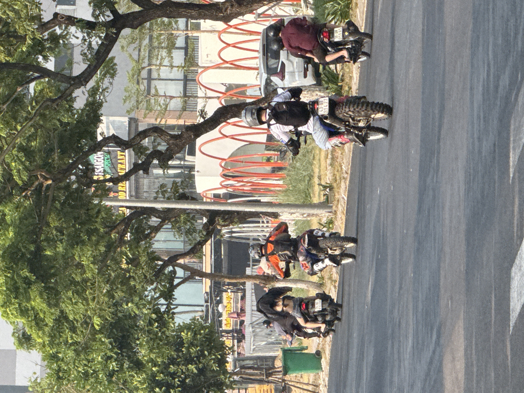 The image shows a street scene in Đà Nẵng, Vietnam. On the right is a gray asphalt road. Several motorcycles are driving on the road. The first motorcycle has a rider in a maroon top. The second motorcycle has a rider with a helmet, white shirt, and black backpack. The third motorcycle has a rider in orange and black. The last motorcycle has a rider in black. On the left side of the street, there is a sidewalk with green bins and trees. Behind the sidewalk, buildings are visible with signs, including one that says "SAIGON Restaurant". In the background is a white structure with red, curved lines.