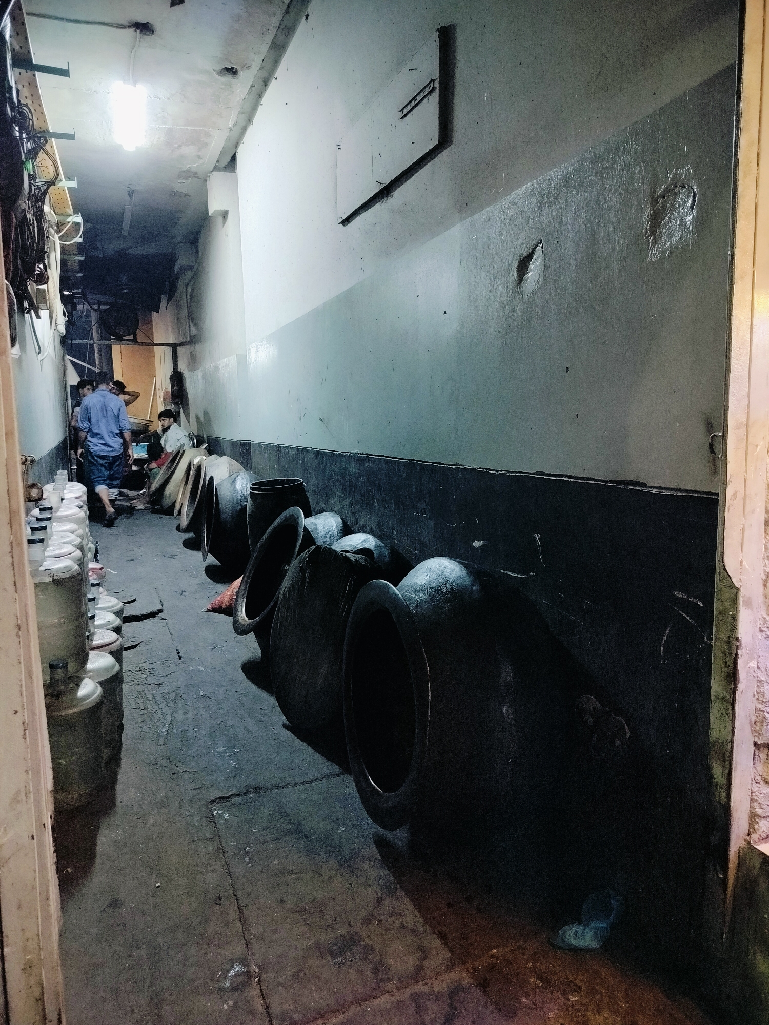 In a narrow, dimly lit alleyway in Dhaka, Bangladesh, a row of large, dark, earthenware pots are lined up against a grey and black striped wall. Some of these pots are tilted, revealing their wide openings. To the left of the pots, several large, transparent water containers with white lids are stacked. In the background, three men are visible. One man, wearing a blue shirt and shorts, walks away from the viewer. Another man sits near the pots, and a third man stands behind him. The environment appears to be an indoor space, possibly a storage area or a workshop, with a concrete floor. Electrical wiring and conduits are visible along the ceiling and upper walls. The overall impression is one of a functional, utilitarian space, likely associated with some form of traditional craft or trade.