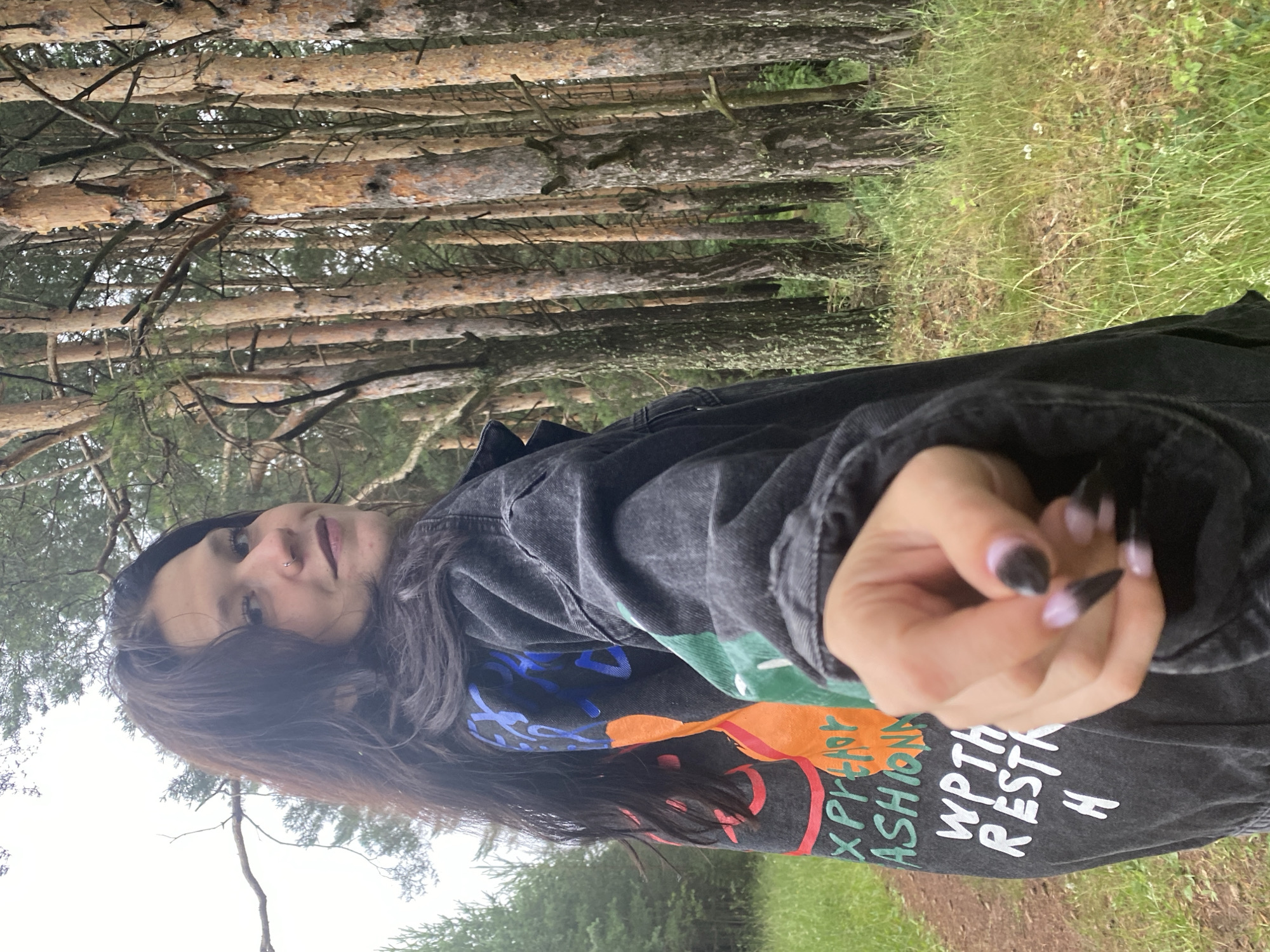 A young woman with long, dark hair is pictured in a forest. She is wearing a dark jacket with a colorful graphic print and the words "WPTH RESTH H" and "XPPEAP ASHIONX" visible on it. Her nails are painted black and light purple. The woman is looking towards the camera, with a slight smile. Her right hand is extended towards the viewer. The background consists of tall pine trees and green grass, suggesting an outdoor setting in a forest. The lighting appears to be natural, likely from daylight, with overcast conditions indicated by the soft light and muted colors. The orientation of the image is landscape, rotated 90 degrees clockwise. The location is identified as Фоки, Russia.