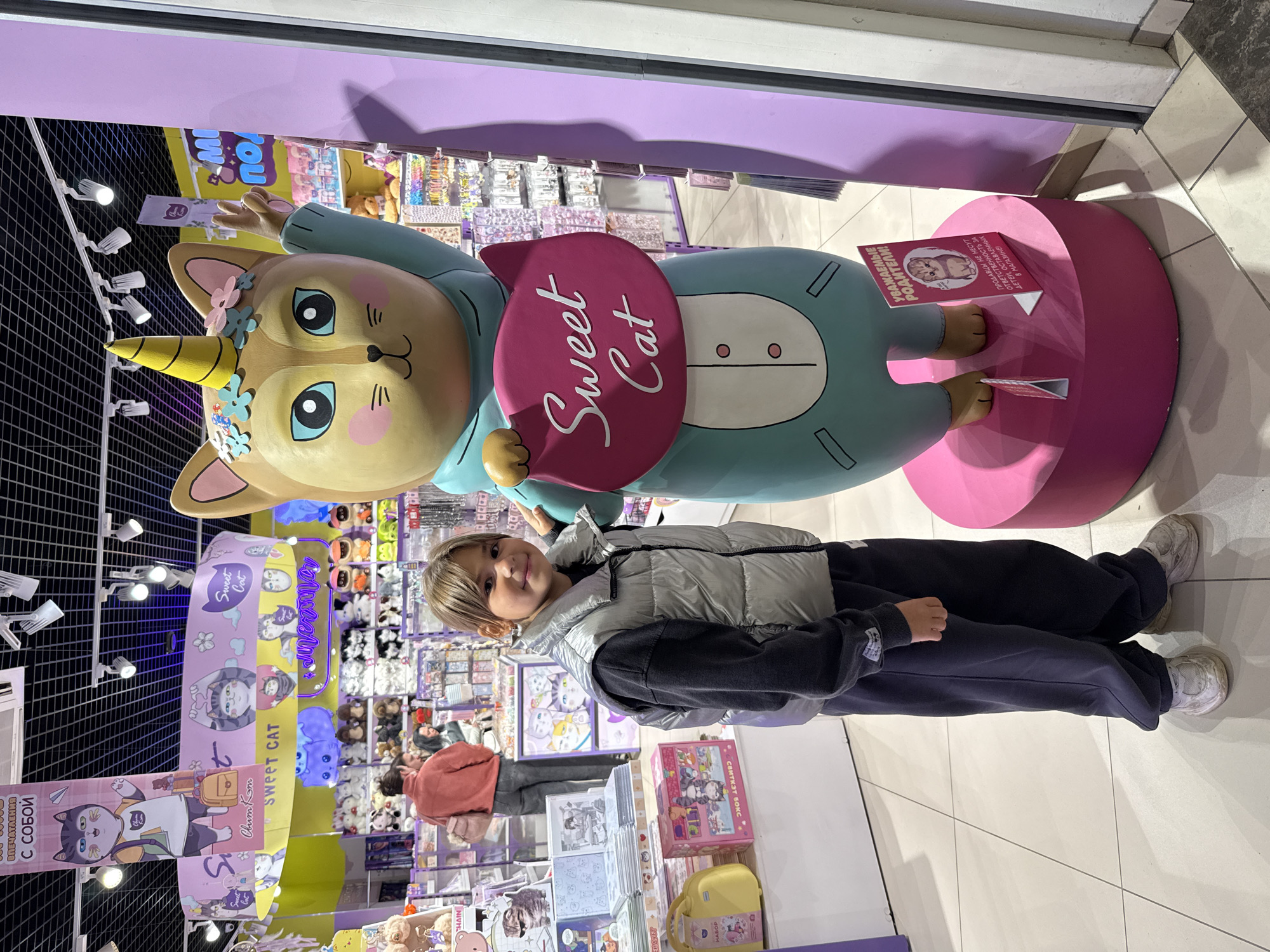 The image captures a young child standing next to a large, cartoonish cat statue with a unicorn horn. The child, with short brown hair and wearing a silver puffer vest over a dark gray sweatshirt and dark gray sweatpants, stands on a tiled floor. The statue is a light brown cat with large blue eyes, pink cheeks, and a yellow horn, wearing a turquoise outfit. It holds a pink heart-shaped sign with "Sweet Cat" written in white script. The scene appears to be inside a brightly lit retail store, likely a toy or gift shop, with shelves of merchandise visible in the background. Banners with cat imagery hang from the ceiling. A small pink sign with Russian text and a picture of a kitten is placed at the base of the statue. The overall impression is of a playful and child-friendly environment.