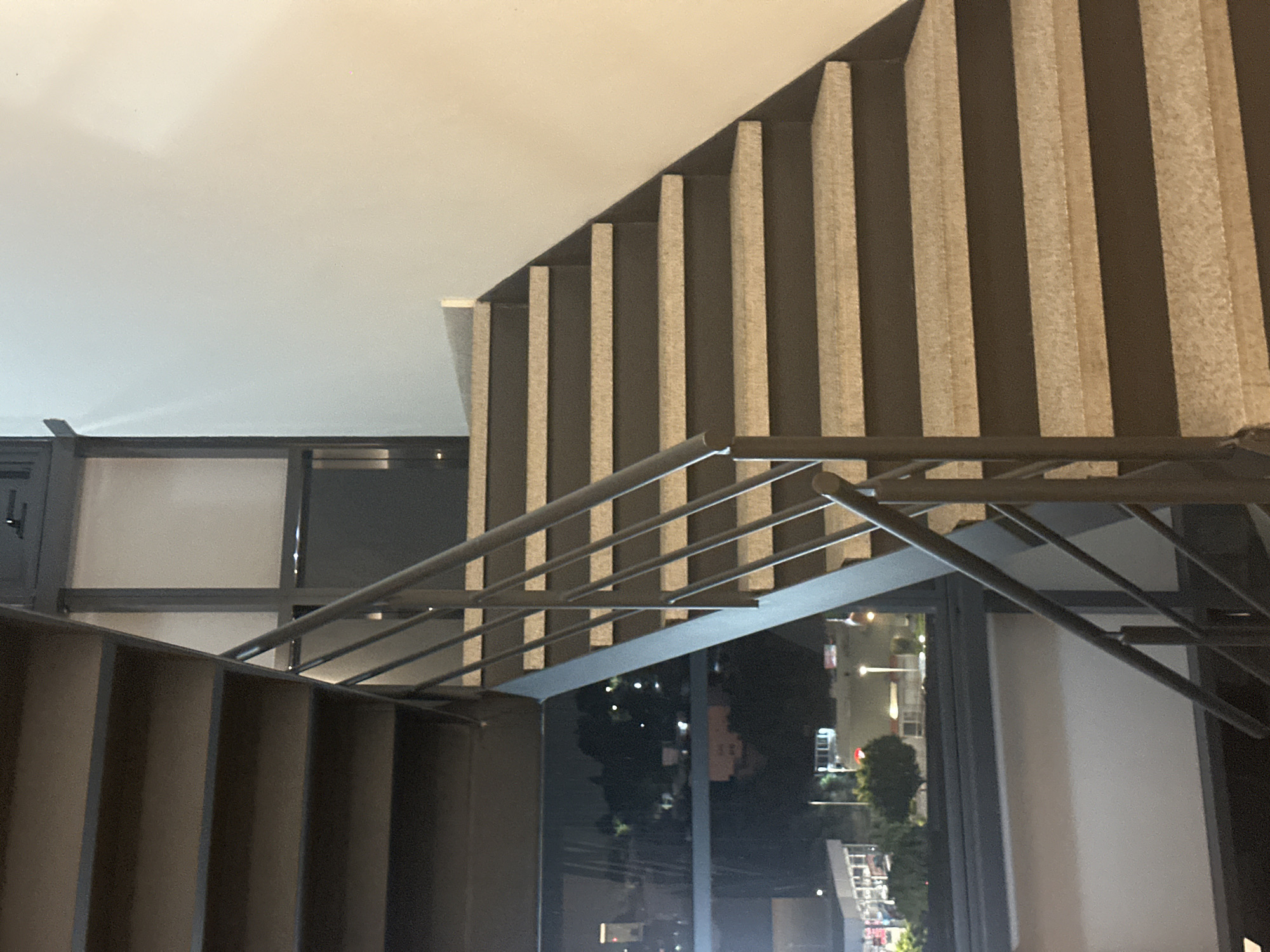The image displays an interior view of a modern building, primarily focusing on a staircase and an expansive window looking out onto a nighttime urban landscape in Huixquilucan de Degollado, Mexico.
The staircase is characterized by a sleek, contemporary design. The lower section, positioned in the foreground, appears to have dark, solid steps and risers. As the staircase ascends, its side wall features a distinctive pattern: light-colored, vertically oriented rectangular panels set against a darker wall. These panels vary in height, creating a visually textured and stepped appearance that complements the incline of the stairs. A minimalist metal railing, likely dark gray or black, with horizontal balusters, runs alongside the staircase, providing a clean architectural line.
To the right of the staircase, a large window reveals the cityscape at night. The scene outside is dominated by darkness, illuminated by numerous artificial lights from buildings, streetlights, and what appear to be distant vehicles, hinting at a lively urban environment. Some dark silhouettes of trees are visible amidst the glow. The time of day is clearly night.
No individuals are visible within the frame, and no specific activity is taking place; the image captures a static architectural moment. The interior spaces are subtly lit, likely by hidden fixtures, creating a gentle contrast with the brighter, artificial illumination of the outside world. The composition suggests an elevated viewpoint within a building, looking down the stairs and across the nighttime city. There is no visible text in the image.