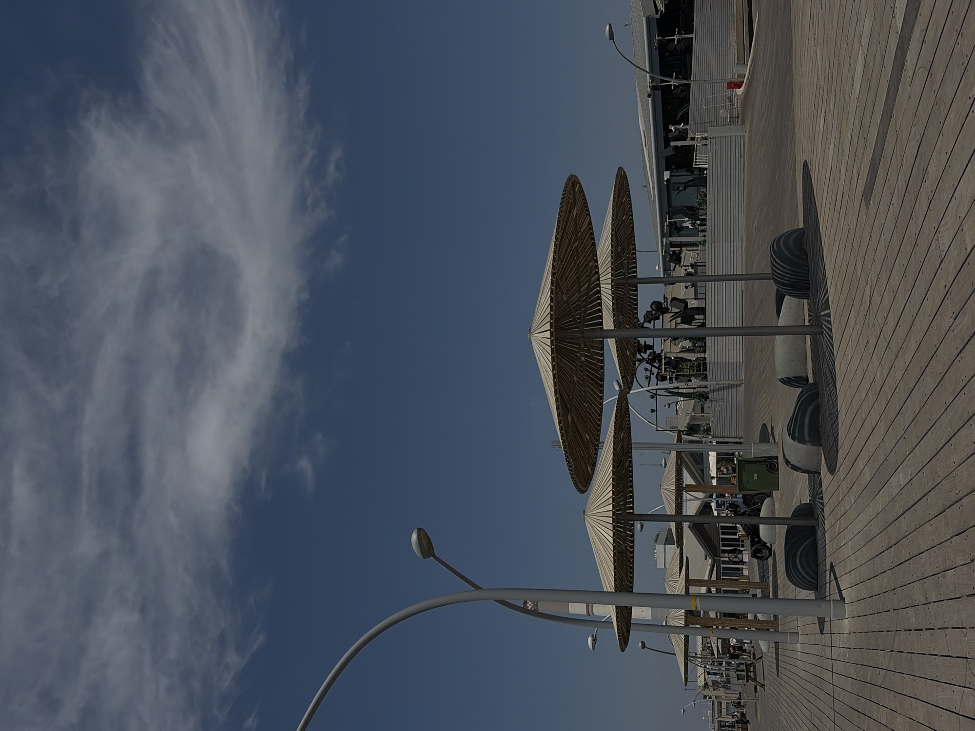 This media file shows an outdoor scene, likely a promenade or boardwalk, under a clear blue sky with wispy white clouds. The perspective is a low-angle shot looking upwards, with the image rotated 90 degrees counter-clockwise.
The primary subjects are a series of large, conical straw umbrellas providing shade over a wooden deck. The umbrellas are supported by white poles. The wooden deck is made of planks and appears to be a walkway. In the background, there are structures with buildings and some seating areas, suggesting a public space like a beach resort or a waterfront area. There are also streetlights visible along the walkway.
The scene appears to be during the daytime, given the bright sunlight casting shadows from the umbrellas. The weather seems pleasant, with a clear sky. There are no overt activities or events depicted, suggesting a static scene of an available public space.
Notable details include the textured straw of the umbrellas, the parallel lines of the wooden planks on the deck, and the curved design of the lampposts. The image is taken from a perspective that emphasizes the scale of the umbrellas against the sky. There is a green trash bin visible in the background.
The location context provided is Tel-Aviv, Israel. This might imply a coastal setting, which aligns with the presence of umbrellas and a boardwalk. No text is readily visible in the image. The overall mood of the image is one of tranquility and a relaxed atmosphere.