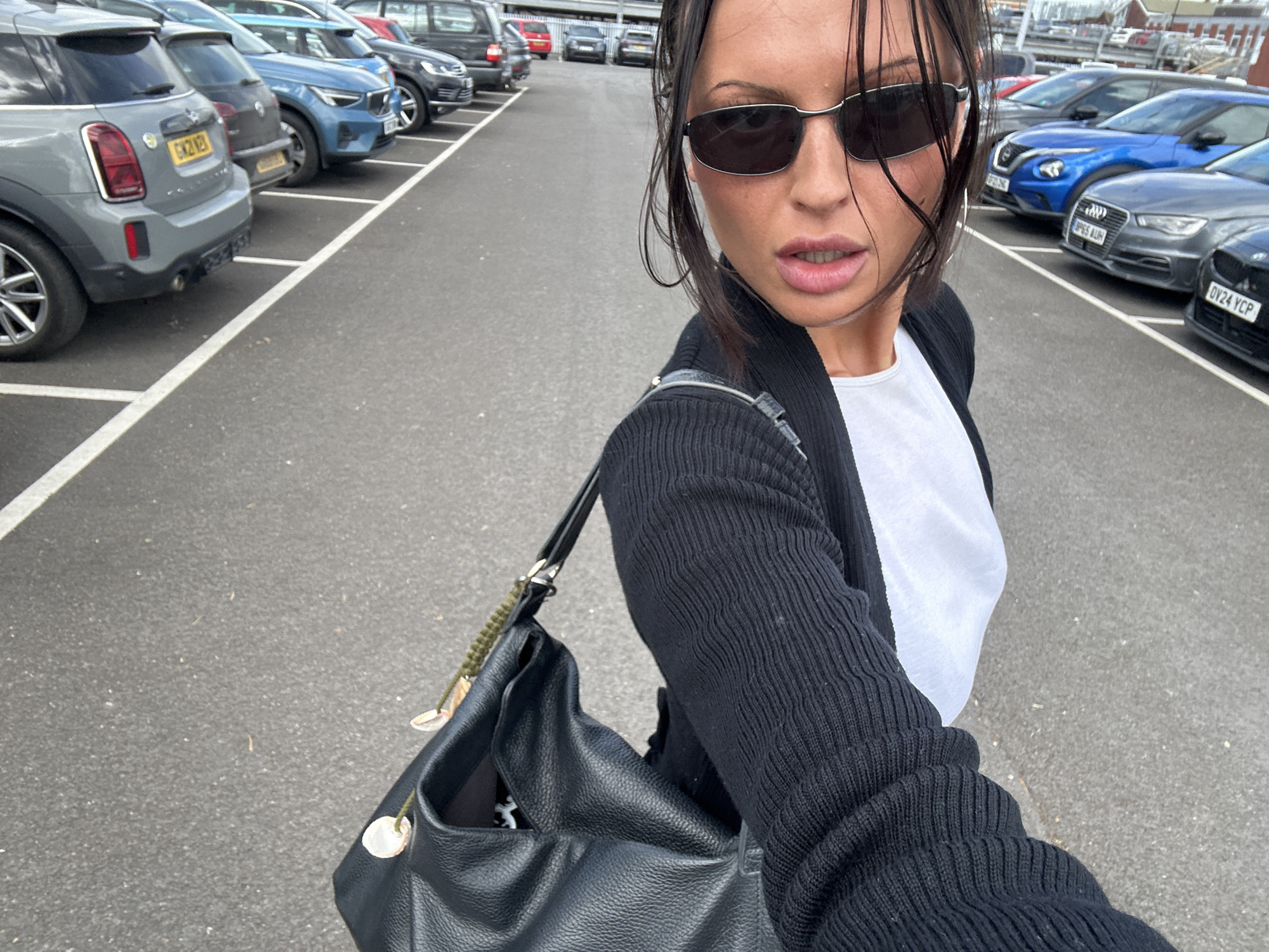 The image captures a person, likely female, wearing sunglasses and a black cardigan over a white shirt, positioned in a parking lot. The individual has a black leather bag slung over her shoulder. Cars are parked in orderly rows on both sides of the asphalt lot, marked with white lines. License plates indicate a United Kingdom location. Based on other visual evidence, this is the parking lot for Sainsbury's in Tunbridge Wells, UK.