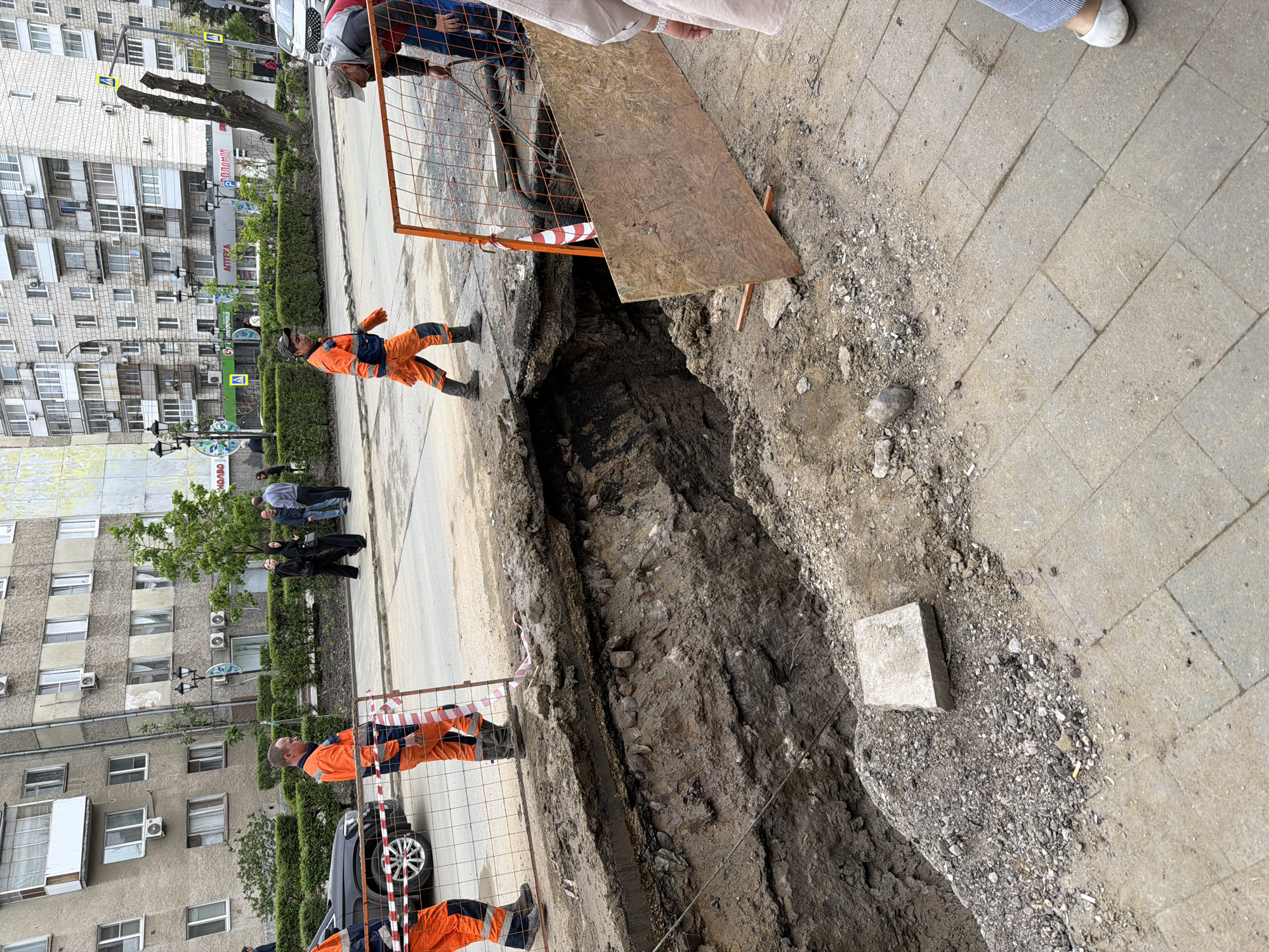 The image depicts a large, open trench in a city street, likely for utility work. It appears to be in Saratov, Russia, based on signage in the background.
**Environment:** The scene is an urban setting with multi-story residential buildings lining the street. The street has sidewalks and a paved road.
**People:**
* Several workers wearing orange jumpsuits and boots are present, positioned near the trench.
* Other people are visible in the background, including those observing the trench.
**Objects:**
* A large trench is the focal point, running alongside the street.
* Safety barriers, including orange fencing and a wooden board, are placed around the trench to prevent accidents.
* A car is parked near the trench.
* A concrete block is near the trench.
**Actions/Interactions:**
* Workers are present near the trench, possibly engaged in ongoing work.
* Bystanders are observing the scene.
* Safety barriers have been erected to cordon off the hazardous area.
**Details:** The trench exposes layers of soil and possible utility lines. The presence of workers in safety gear suggests an active construction or repair site.