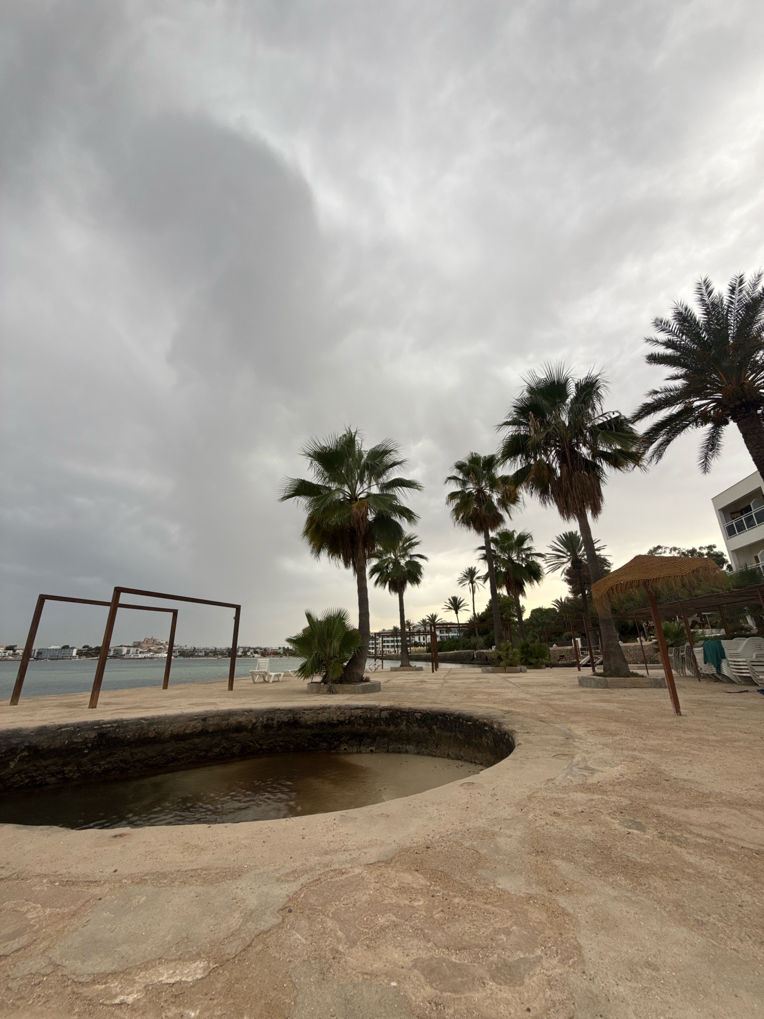 The image depicts a cloudy day on a beach in Eivissa, Spain. In the foreground, there is a circular, shallow pool of dark water with a sandy bottom, surrounded by a textured, light-colored concrete surface. To the left, two tall, rusty metal rectangular frames stand on a sandy area overlooking the water. Beyond this, a calm body of water is visible, with buildings and a hill with structures on it in the distance. The middle ground is dominated by several tall palm trees, some with dried fronds. To the right, there is a white building with balconies, a thatched umbrella, and stacks of white lounge chairs. The sky is overcast with heavy gray clouds. There are no people visible in the frame.