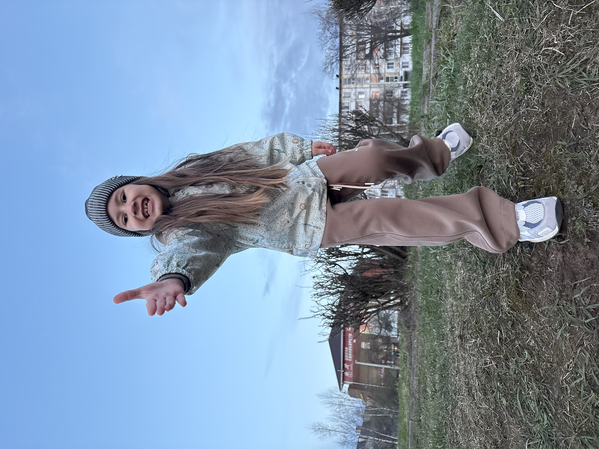 A young girl, seen from a low-angle perspective, stands outdoors, filling the majority of the frame. She has long, straight brown hair and is wearing a striped beanie, a light green long-sleeved jacket with a subtle pattern, loose-fitting brown pants, and white and grey athletic sneakers. She is smiling broadly, revealing her teeth, and appears joyful and playful. Her left arm is extended towards the camera with an open palm, as if reaching out or gesturing an invitation. Her right arm is bent, and she holds a small, light-colored rectangular object, possibly a phone or toy.
The background features a clear blue sky with some scattered white clouds above the girl. Below her feet is a patch of ground covered with dry, sparse green and brown grass. In the mid-ground, behind the girl, there are several bare trees, suggesting a cooler season like early spring or late autumn. Further back, a multi-story residential building with numerous windows is visible, along with another structure featuring a red facade. On this red building, Cyrillic text is visible, which reads "ЦЕНЫ СНИЖЕНЫ", translating to "PRICES REDUCED". The lighting indicates daytime, possibly late afternoon, with soft, diffused light. The overall mood of the image is cheerful and engaging.