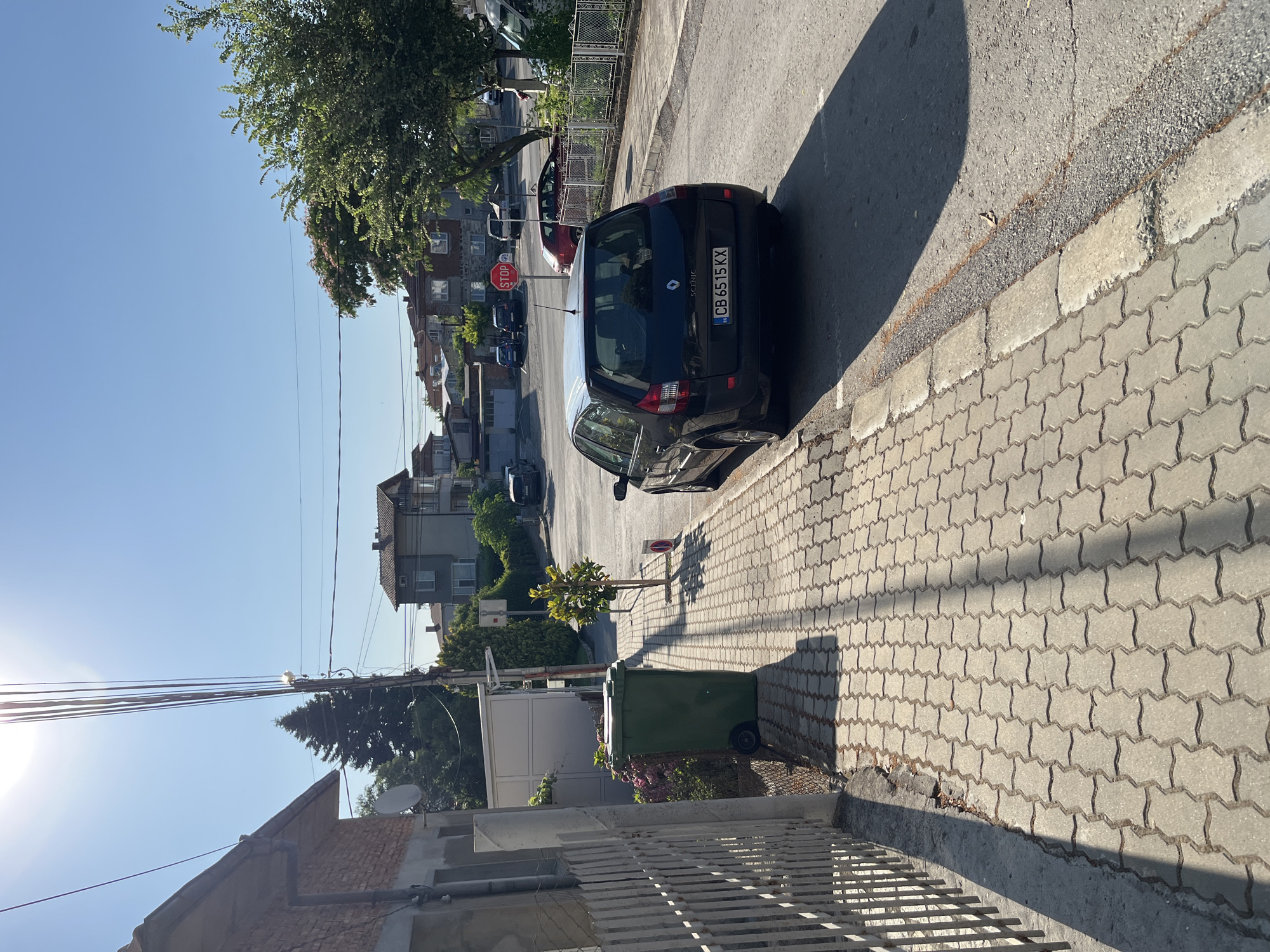 This image captures a sun-drenched street scene in Obzor, Bulgaria. The perspective is from a sidewalk, looking down a gently curving road. On the left, a weathered brick building with a grey facade and a white fence runs alongside the sidewalk. Adjacent to the building is a large green garbage bin. The sidewalk itself is paved with interlocking grey bricks.
The road is paved with asphalt and has a curb. Parked on the right side of the road is a black Renault Scenic car with Bulgarian license plates "CB 6515 KX". Further down the road, other vehicles are visible, including a red car and a white van, along with houses and trees. A red STOP sign is posted on a pole at an intersection in the distance. The sky is clear and blue, and the sun is high, casting distinct shadows across the scene. The environment appears to be a residential area. There are no people visible in the image.
