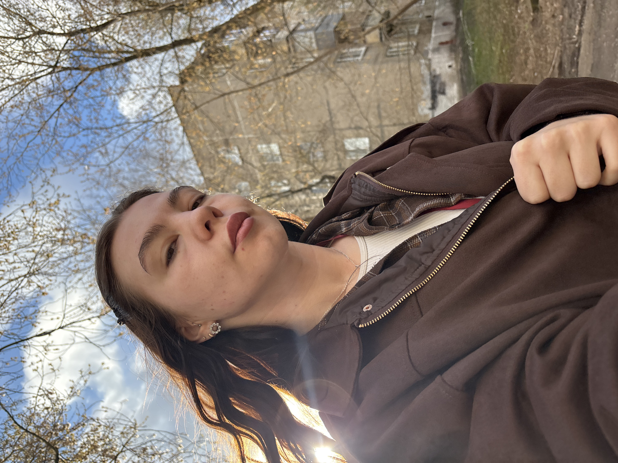 This low-angle, rotated selfie captures a young person, likely female, looking directly at the camera with a slightly pensive or pouty expression. Her long, dark brown hair frames her face, with strands falling towards the bottom of the frame. She is wearing a brown jacket with a prominent brass-colored zipper, layered over a white ribbed top and a brown plaid shirt, visible through the open jacket. In her left ear, she sports two earrings: a small hoop and a decorative, circular studded piece. Her left hand is visible, lightly gripping the jacket fabric.
The background is an upward view of a bright blue sky with scattered white clouds, interspersed with bare tree branches, suggesting late autumn, winter, or early spring. Through the branches, a multi-story, light-colored apartment building with several windows is visible in the distance. A strong lens flare emanating from the lower left part of the image indicates the sun is low in the sky, suggesting either morning or late afternoon. The scene is set outdoors in Perm, Russia, likely within a residential area. No text is visible in the media.