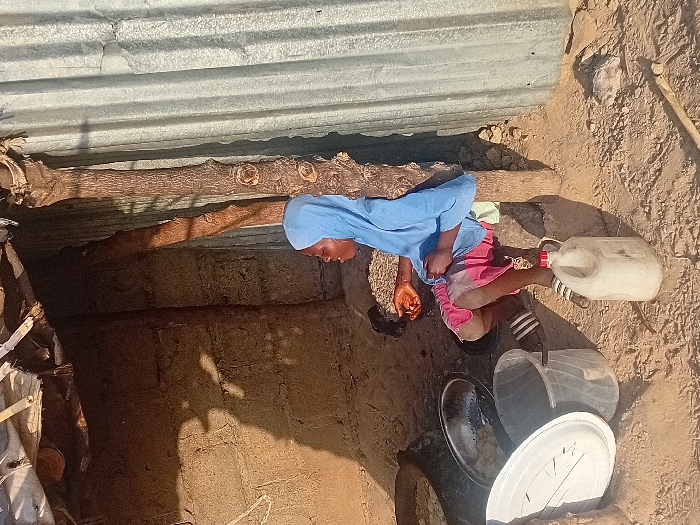 A young person in a light blue head covering and shirt, and pink shorts, is squatting inside a rustic structure with a corrugated metal roof supported by wooden beams. Next to them, there is a white plastic jug, black and white striped sandals, and a stack of bowls and a lid. The floor is dirt, and the walls appear to be made of mud brick. The lighting is dim, highlighting the interior's simple and possibly impoverished conditions.