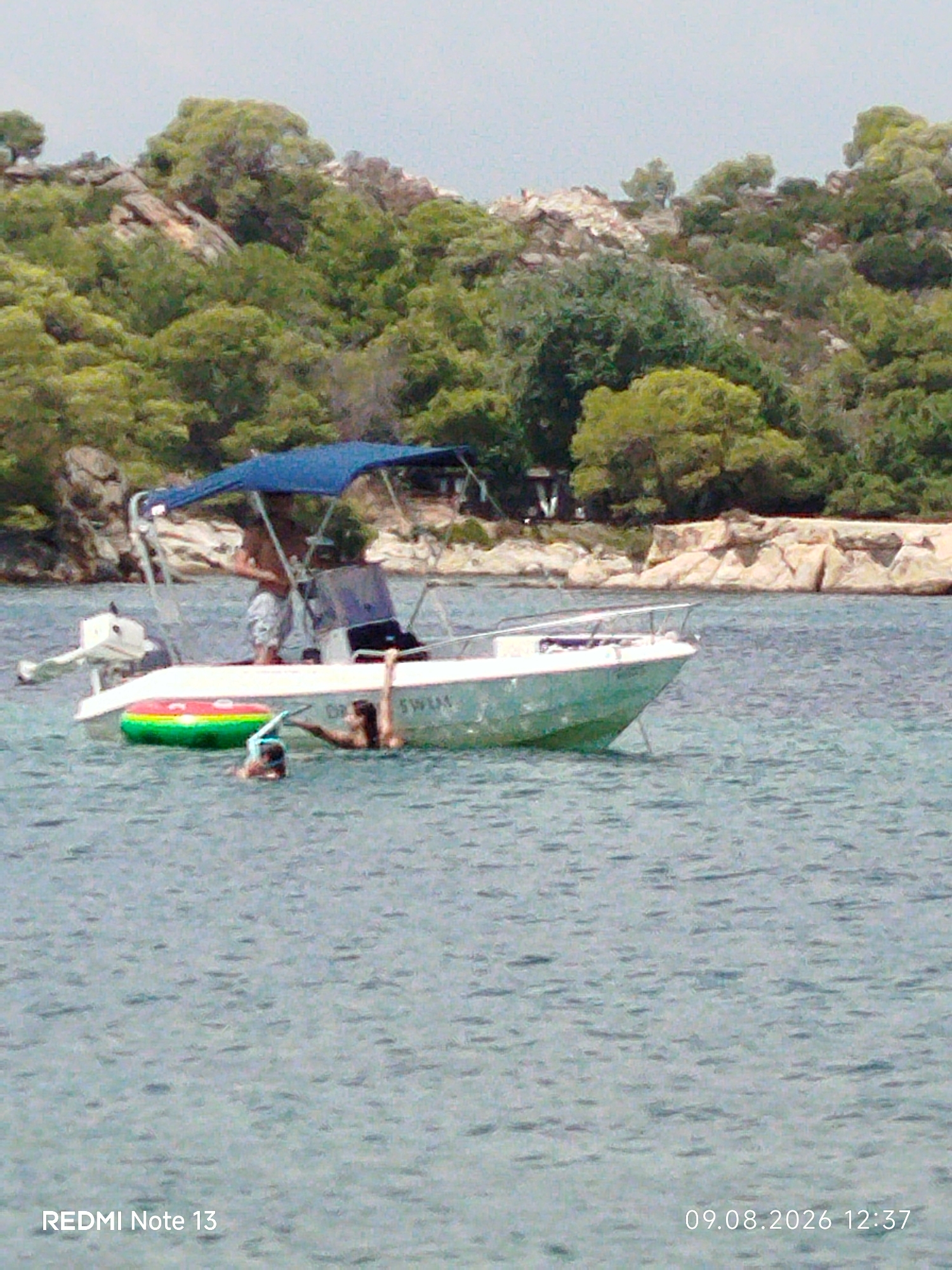 A small white motorboat with a blue canopy is moored in the clear, blue waters off a rocky, tree-covered coastline. The boat has an outboard motor at the stern and a person standing in it. Two people are in the water near the boat; one is wearing a snorkel mask, and the other is partially submerged. A brightly colored inflatable ring floats beside the boat. The background shows a lush green hillside dotted with rocks and trees, with a hazy sky above. The image appears to have been taken in Sithonia, Greece, given the natural landscape and clear waters.