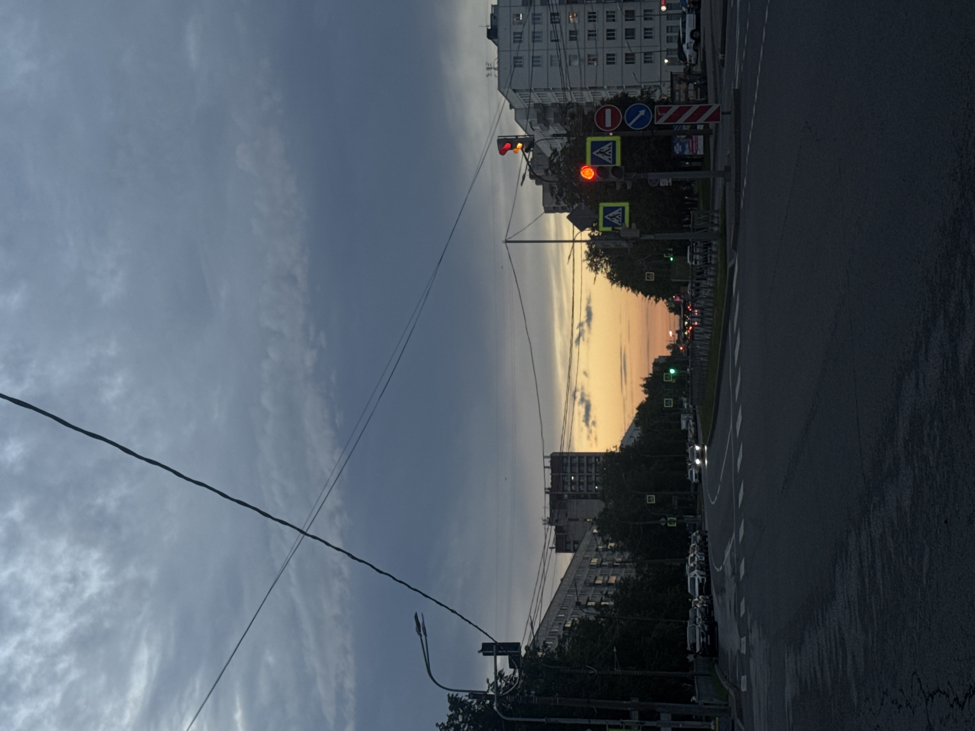This is a medium-shot photograph of a street scene in Ladozhskoe Ozero, Russia, taken at dusk. The sky is filled with heavy, dark clouds that are beginning to break, revealing a sliver of orange and yellow sunset on the horizon. Power lines crisscross the sky. A street light with two fixtures is visible on the left, and a traffic light on the right displays a red signal. Several cars are parked along the left side of the road, and a few cars are driving on the road, their headlights illuminated. Pedestrian crossing signs are present at the intersection. Tall apartment buildings line the street, with lights visible in some windows. Trees are interspersed along the roadside. The road surface appears wet and slightly cracked.