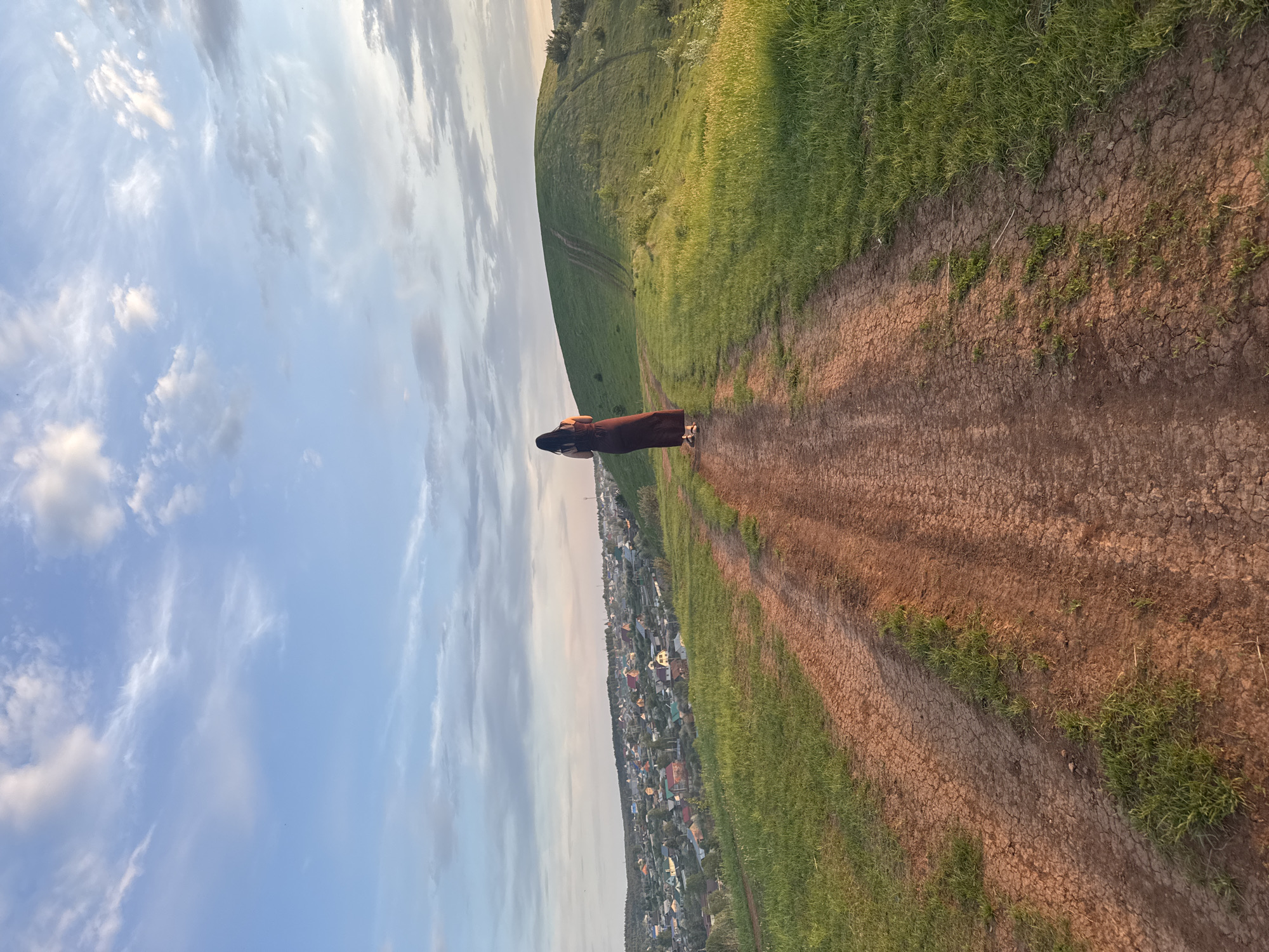 A long-shot captures a person standing in the middle of a dirt path, alongside a grassy hill, with a rural village in the background under a partly cloudy sky. The person is wearing a long, dark brown dress and has long, dark hair. The dirt path, predominantly brown, shows signs of cracking and sparse green vegetation. The grassy hill is vibrant green. In the background, the rural village, presumed to be in Ольховка, Russia, consists of numerous buildings and houses. The sky is blue with scattered white clouds.