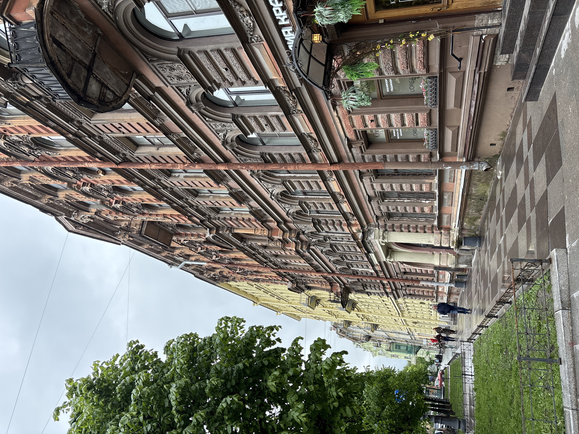 The image captures a street view in Saint Petersburg, Russia. The shot is taken from a low angle, looking upwards towards ornate, multi-story buildings. The buildings exhibit a variety of architectural details, including windows, arches, moldings, and decorative elements. One building features a sign with Cyrillic text. A large tree with green foliage is visible in the lower portion of the frame. Pedestrians can be seen walking along the sidewalk in the distance. The sidewalk is paved with a checkered pattern, and a low metal fence separates the walkway from a strip of grass. The sky is overcast, suggesting a cloudy day.