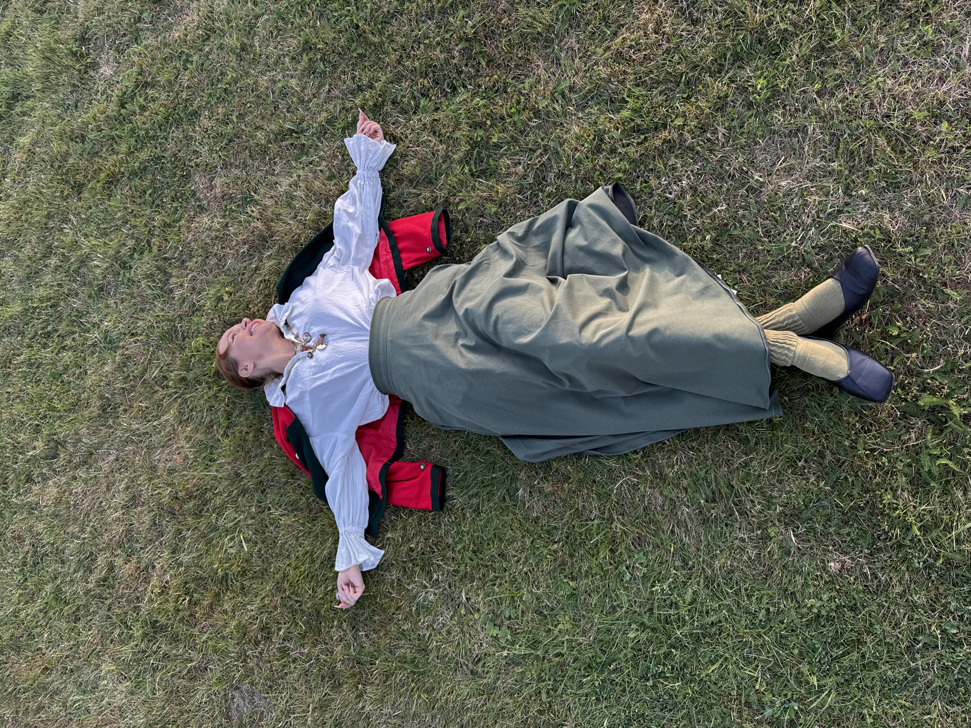 A person lies on their back in a grassy field, appearing to be in the city of Arkhangel'skoe, Russia. They are dressed in a white ruffled blouse, a red jacket with dark green trim, a long olive green skirt, tan socks, and dark shoes. Their arms are outstretched to the sides, and their head is tilted back, eyes closed, with a slight smile on their face. The person's red hair is visible. The environment is a sunlit field of green and brown grass.