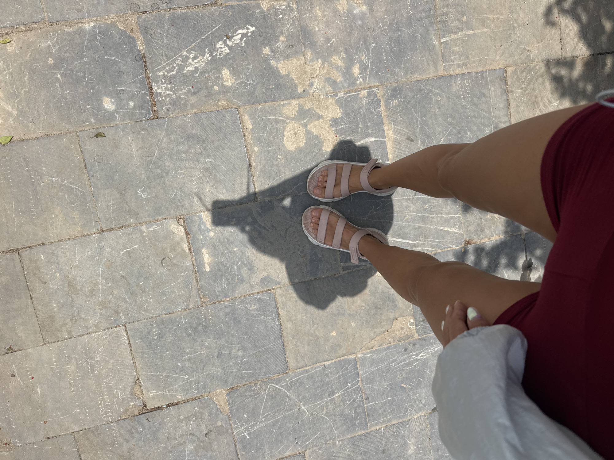 An eye-level shot shows a section of a paved area with a person standing on it. The paving consists of gray, rectangular stones with visible scratches and imperfections. The person's legs, clad in light pink sandals, are positioned in the center of the frame. They are wearing maroon shorts. A portion of a white garment, possibly a jacket or bag, is visible in the lower right corner. The location is Mèo Vạc Commune, Vietnam.