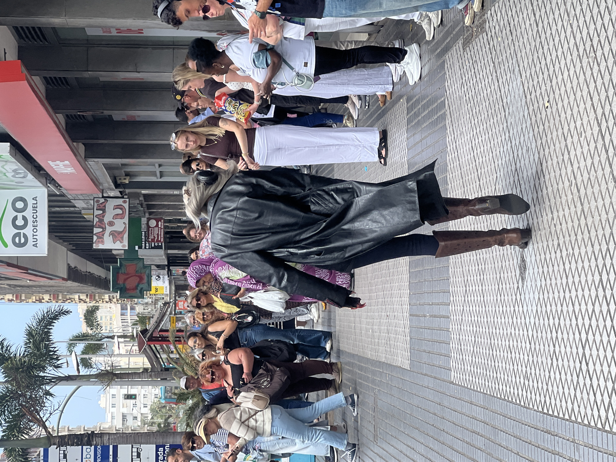 The image depicts a vibrant street scene in Las Palmas, Spain, during daylight hours under a clear, sunny sky. The perspective is from behind a person wearing a black leather coat, dark trousers, and brown boots, walking on a textured paved sidewalk.
A diverse crowd of people, mostly adults, are walking and standing along the pedestrian street. They are dressed in casual, warm-weather attire, including t-shirts, blouses, jeans, and wide-leg pants. Many individuals are carrying handbags or shoulder bags, and some wear sunglasses. One person is visible holding a snack bag with the text "TRIX" and "CARN..." prominently displayed.
The street is flanked by multi-story commercial buildings, displaying various shop signs. Palm trees are visible further down the street, confirming the warm climate. A traffic light can also be seen. The sidewalk features a distinct diamond pattern on one side and grooved horizontal lines on the other.
Visible text on signs includes "eco AUTOESCUELA" (a green sign), "BAZARO" (a red sign), and "PARA LLEVAR" (likely indicating a take-away food service). Other partial texts such as "CHA upsilo," "PROVISION," and "rada ioni" are also present on various street signs and advertisements. The overall atmosphere appears relaxed, consistent with a typical day in a bustling city.
