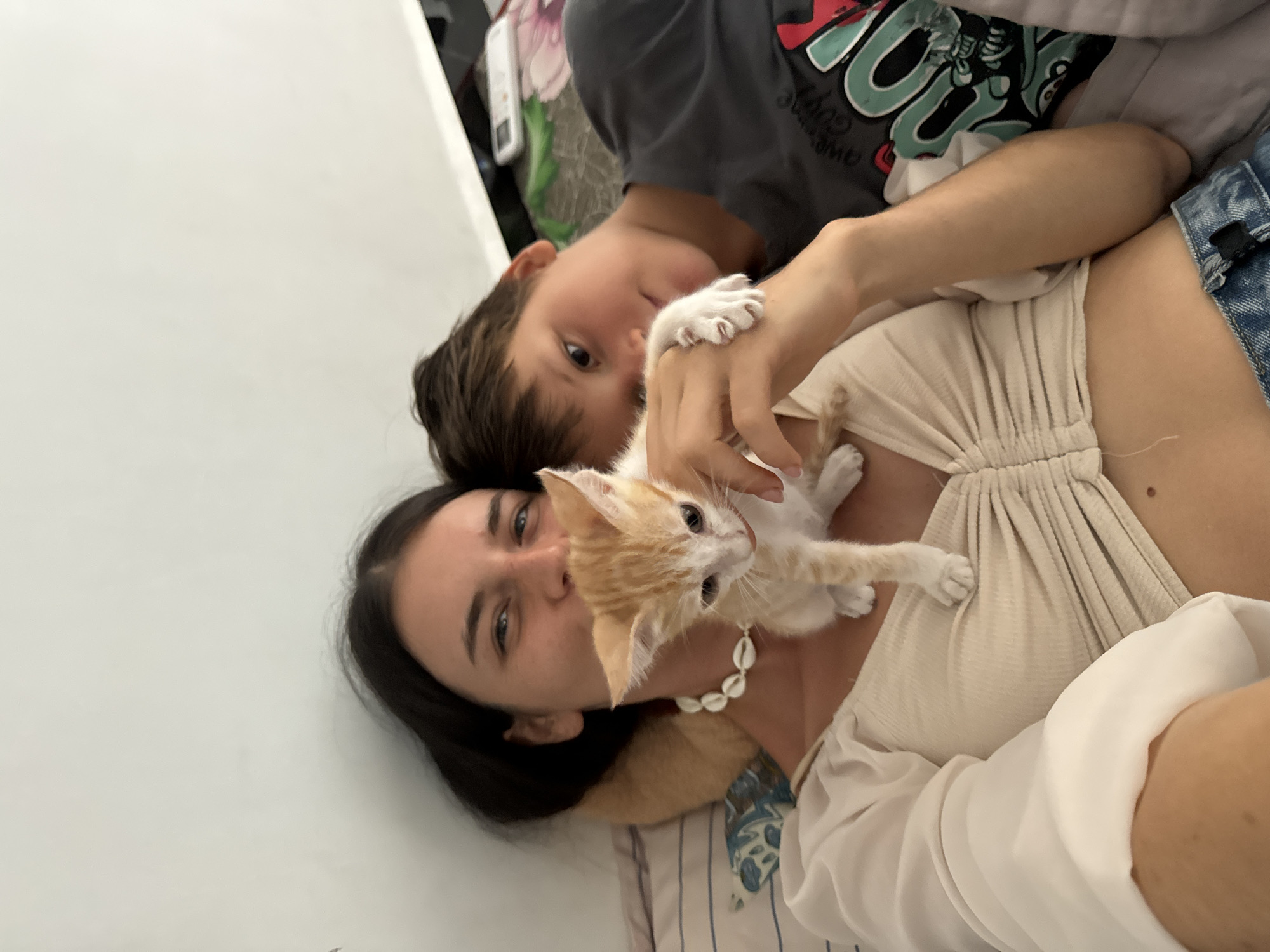 A young woman and a boy lie in bed with a small ginger and white kitten. The woman is in the foreground, wearing a light-colored, ruched top and denim shorts. She is smiling and holding the kitten, which is perched on her chest. The boy is behind her, to the right, wearing a dark t-shirt. The kitten is looking to the left. The environment appears to be a bedroom in a tropical location, possibly Mũi Né, Vietnam, with a white wall and bedding visible. The lighting suggests daylight.