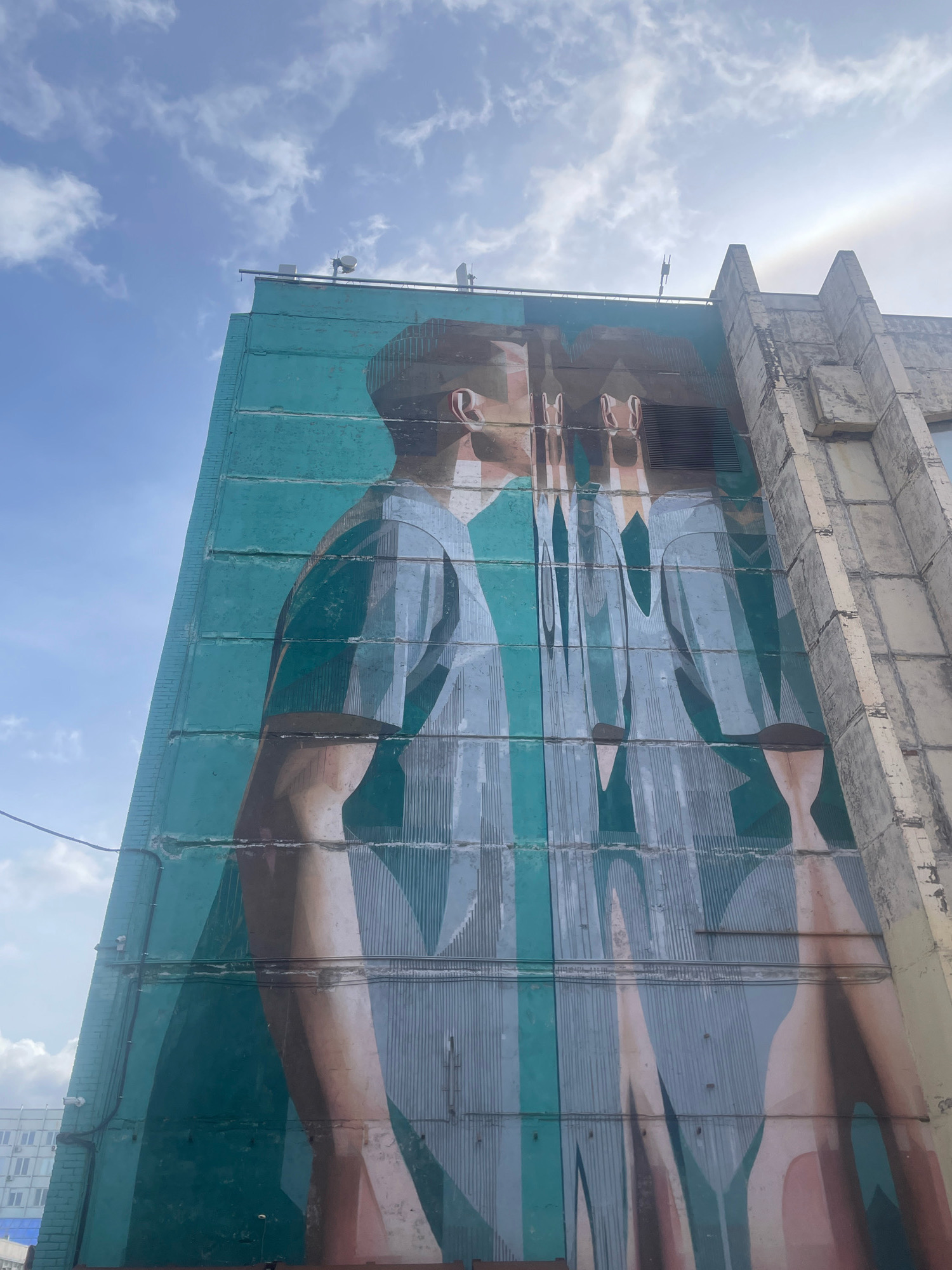 A low-angle, wide shot captures a mural on the side of a Brutalist apartment building, set against a bright, partly cloudy sky. The mural, rendered in shades of teal, grey, and peach, depicts two figures from the shoulders up. The figure on the left is facing right in profile, appearing to be in mid-conversation or about to kiss another figure whose face is obscured and distorted by a rippling effect, suggesting a mirror image or a glitch in perception. The building's facade is segmented by horizontal lines and features some exposed concrete, metal conduits, and ventilation grates. The artwork's style is geometric and abstract, with sharp lines and flat planes of color. The surrounding environment is not clearly visible, but the building suggests an urban setting, possibly in Saint Petersburg, Russia, given the architectural style commonly found in post-Soviet housing complexes. The overall atmosphere is one of urban decay juxtaposed with modern artistic expression.