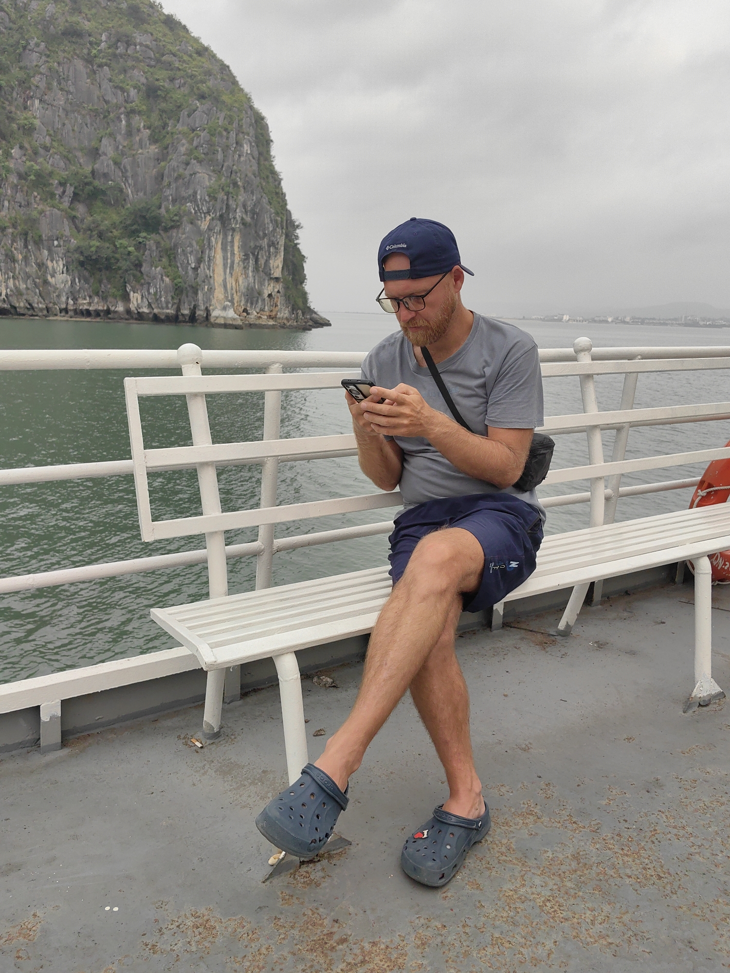 A man is sitting on a white bench on what appears to be a ferry or boat. The man is wearing glasses, a blue baseball cap, a gray t-shirt, blue shorts, and blue Crocs. He is looking at a mobile phone in his hands. He has a reddish beard and a small black bag across his shoulder.
Behind the bench, there is water and a large, rocky, green-covered island. The sky is overcast.
The ground area where the bench sits is gray and shows signs of wear and rust.
The location is Tuan Chau Ward, Vietnam.