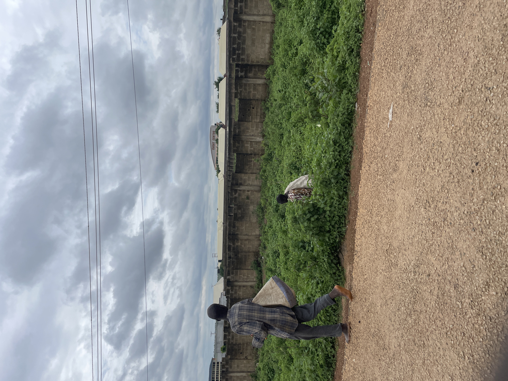 In an outdoor, overcast scene in the city of Unknown, Nigeria, a person walks along a dirt path carrying a large, light-colored bag over their shoulder. They are wearing a plaid shirt and dark pants, and appear to be barefoot. To the right of the path, a person is seen partially obscured by dense green foliage, also appearing to be barefoot and wearing a patterned garment. Behind the foliage is a concrete block wall, beyond which are several buildings, one with signage that reads "EVENT CENTRE." Power lines are visible in the sky.