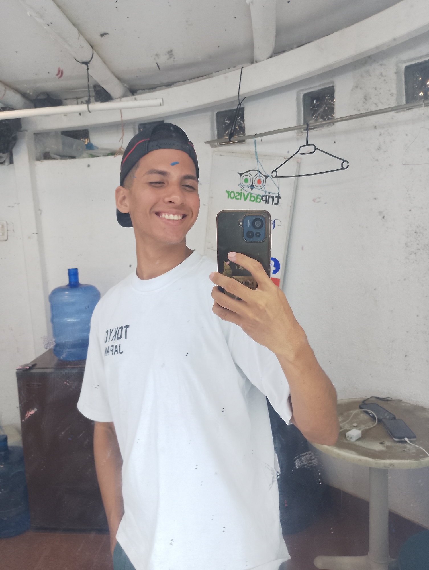 A young man is taking a selfie in a mirror. He is wearing a white t-shirt with "TOKYO JAPAN" printed on it, a black baseball cap with a red stripe, and has a blue mark on his forehead. He's smiling and holding a black smartphone with three camera lenses in his right hand. The mirror reflects a cluttered room. Behind him is a large blue water jug on a brown cabinet. A metal rod runs horizontally along the top of the wall with a coat hanger hanging from it. A small round table stands to the right, with a white charger and two smartphones lying on it. The mirror's reflection shows signs of wear and discoloration, suggesting a humid environment. The location may be Porlamar, Venezuela, based on the presumed context and the overall appearance of the environment.