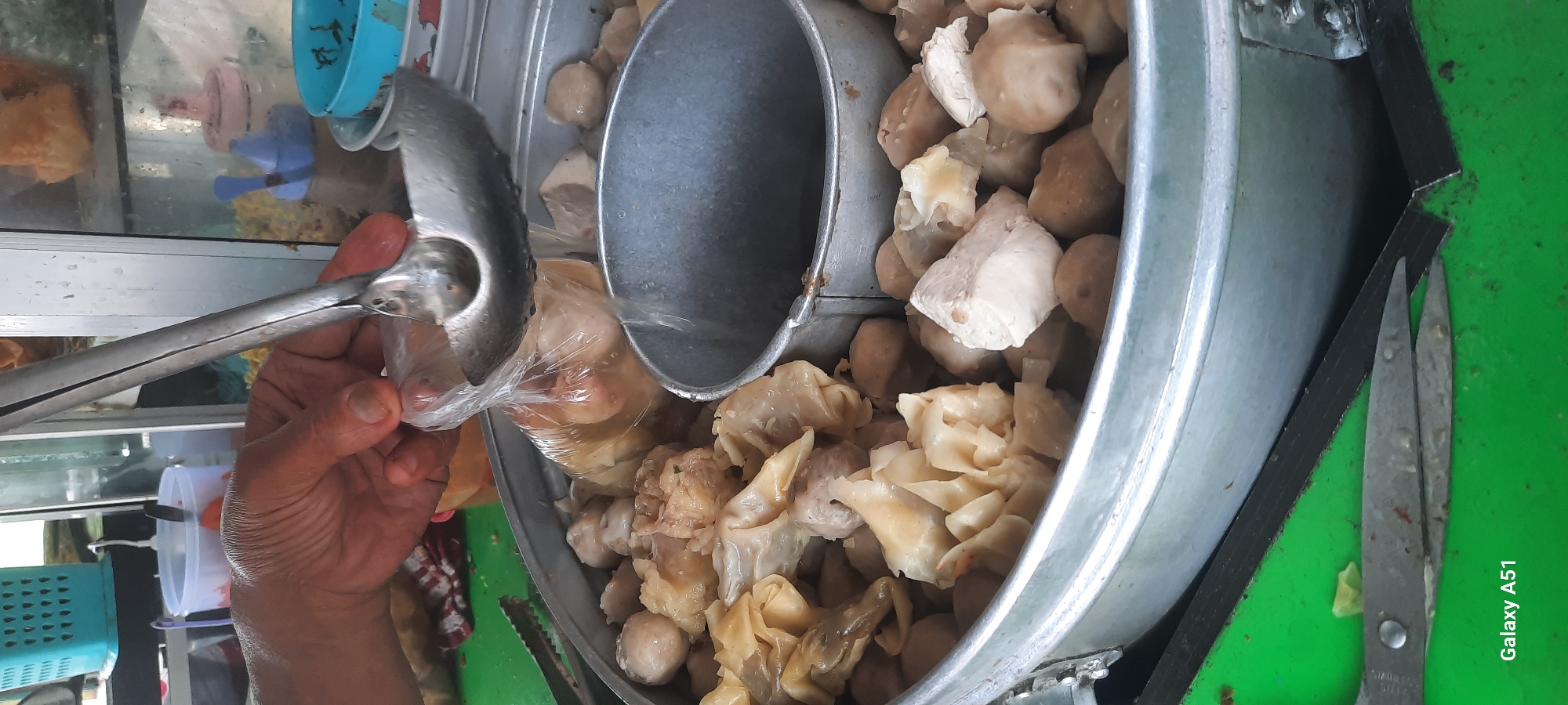 A person's hand, holding a ladle, is scooping food from a large metal pot. The pot is filled with meatballs and some kind of dumplings. In the background, there are containers with condiments and other food items. The scene appears to be a street food stall in Selopuro, Indonesia. The image is taken from a close-up perspective, focusing on the food and the hand preparing it. The lighting is bright, suggesting an outdoor setting during the day.