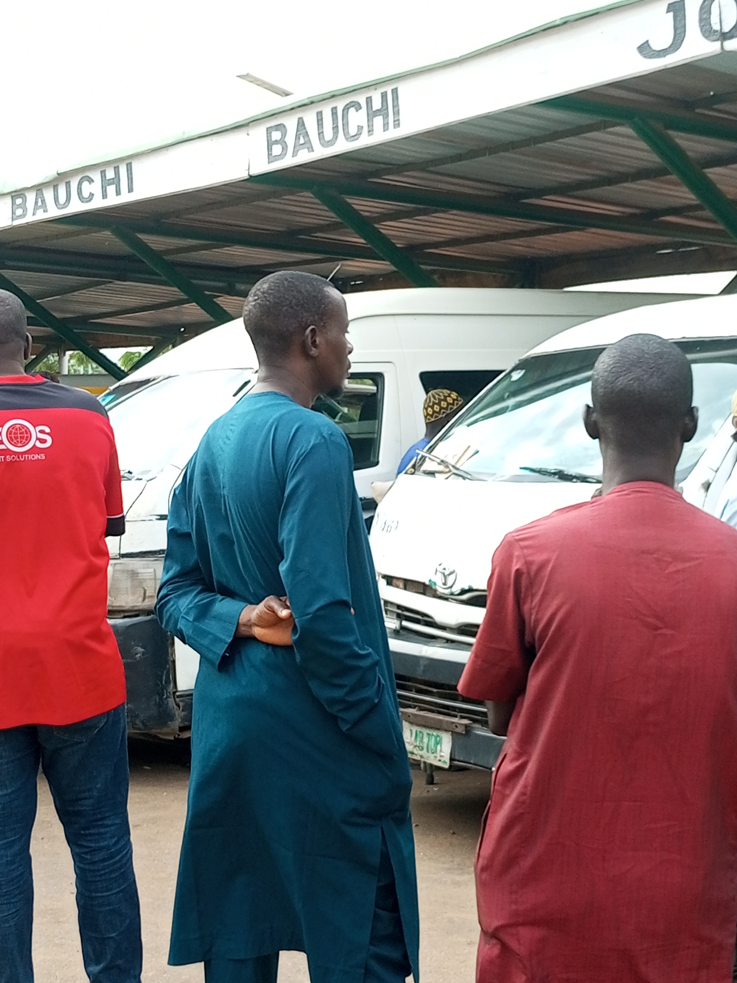 The image shows a street scene in Gombe, Nigeria, at what appears to be a bus station. Several white minibuses are parked under a covered shelter with the word "BAUCHI" and "JO" visible on signs above. Three men are in the foreground. One man on the left wears a red and black t-shirt and blue jeans. In the center, a man in a teal-colored traditional outfit stands with his hands behind his back, facing away from the camera. To the right, another man in a red traditional outfit is also facing away. The ground is dirt, and the overall atmosphere suggests a busy transportation hub.