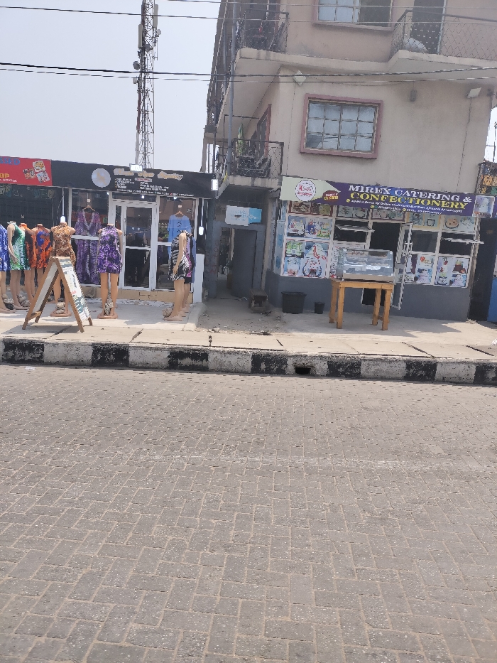 This image depicts a street scene in Alaiyabiagba, Nigeria, observed during daytime. The foreground features a grey paver-block road, laid in a herringbone pattern. A concrete sidewalk, marked by a curb with alternating black and white stripes, separates the road from two adjacent commercial establishments. A small drainage opening is visible in the curb.
On the left, a single-story storefront with a dark facade displays several headless mannequins dressed in vibrant, patterned garments outside its glass entrance. An A-frame sign is positioned on the sidewalk near these mannequins. A sign above the shop entrance includes the text "Unique Collections."
Immediately to the right, a two-story building with a light beige exterior houses "MIRLX CATERING & CONFECTIONERY" on the ground floor. This shop features large display windows covered with various food advertisements and posters. A wooden table supporting an empty glass display case stands directly outside the confectionery. A black bucket rests on the ground nearby. The second floor of this building presents a window with dark framing and a small balcony with a metal railing.
In the upper-left background, a tall, metallic lattice utility pole or telecommunications tower rises above the buildings. The sky is bright and appears clear. No individuals are visible in the scene, and no overt actions or interactions are occurring.