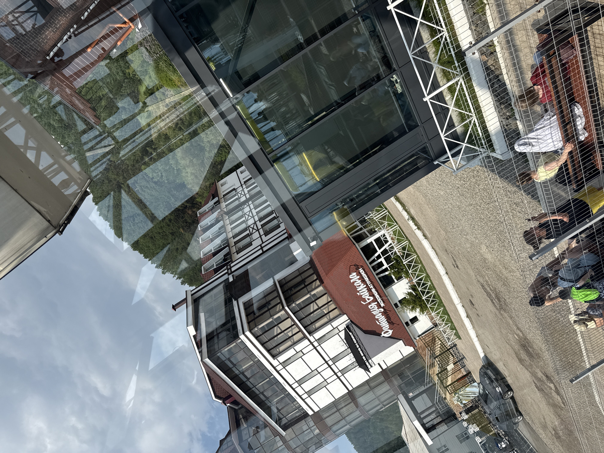 In Listvyanka, Russia, a modern hotel complex with a red-tiled roof and white facade, featuring large glass windows and balconies, is captured. A red sign on one of the buildings reads "Фантазия Байкала" (Fantasy Baikal) along with "Эксклюзив-Аттракцион" (Exclusive Attraction). A black car is parked in the gravel lot in front of the complex, and a white fence with diamond-shaped patterns separates the building from a pathway. In the foreground, several people are visible behind a wire fence, some sitting on a wooden bench. They appear to be tourists, with one person wearing a bright green backpack. The scene is set against a backdrop of lush green forested hills under a cloudy sky. The image appears to be taken from an elevated perspective, possibly from a cable car or another building.