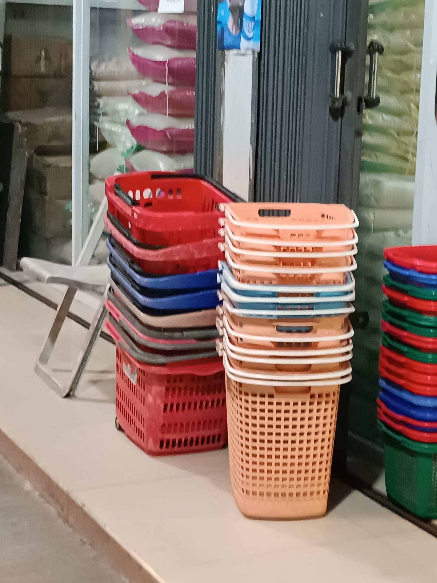 A stack of red, blue, and peach-colored plastic shopping baskets are visible on a tiled floor, with a closed dark metal gate in the background. The baskets are neatly piled, with a few red baskets on the left and a larger stack of peach and blue baskets on the right. To the left of the baskets, a metal folding stool is partially visible. The environment appears to be an indoor retail space, possibly a shop or market. The image is taken from a slightly elevated perspective, looking down at the baskets. The lighting is moderate, casting soft shadows. The background shows rows of merchandise bags, suggesting a retail setting. The image location is identified as Jetis, Indonesia.