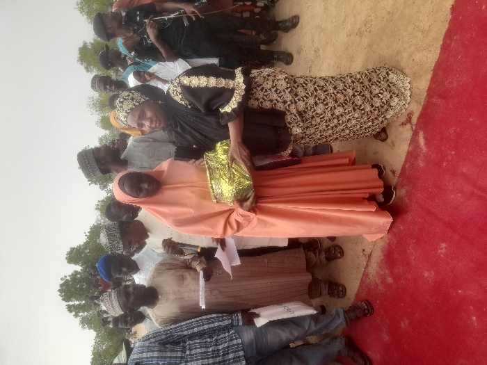 The image shows a group of people gathered outside, presumably in Gusau, Nigeria, based on the provided coordinates. A young woman wearing a peach hijab and a long, peach dress is holding a gold-colored container. She is standing on the right side of a red carpet. A woman wearing a black abaya and a white and gold skirt is standing on the left side of the carpet. She is handing the container to the young woman. The woman in the black abaya is smiling. There are many people standing behind them, many are wearing white robes with caps. The people in the background are not clearly visible. The ground is light brown. There are trees in the background. The sky is overcast. The image appears to be a candid snapshot, possibly captured at an event or ceremony.