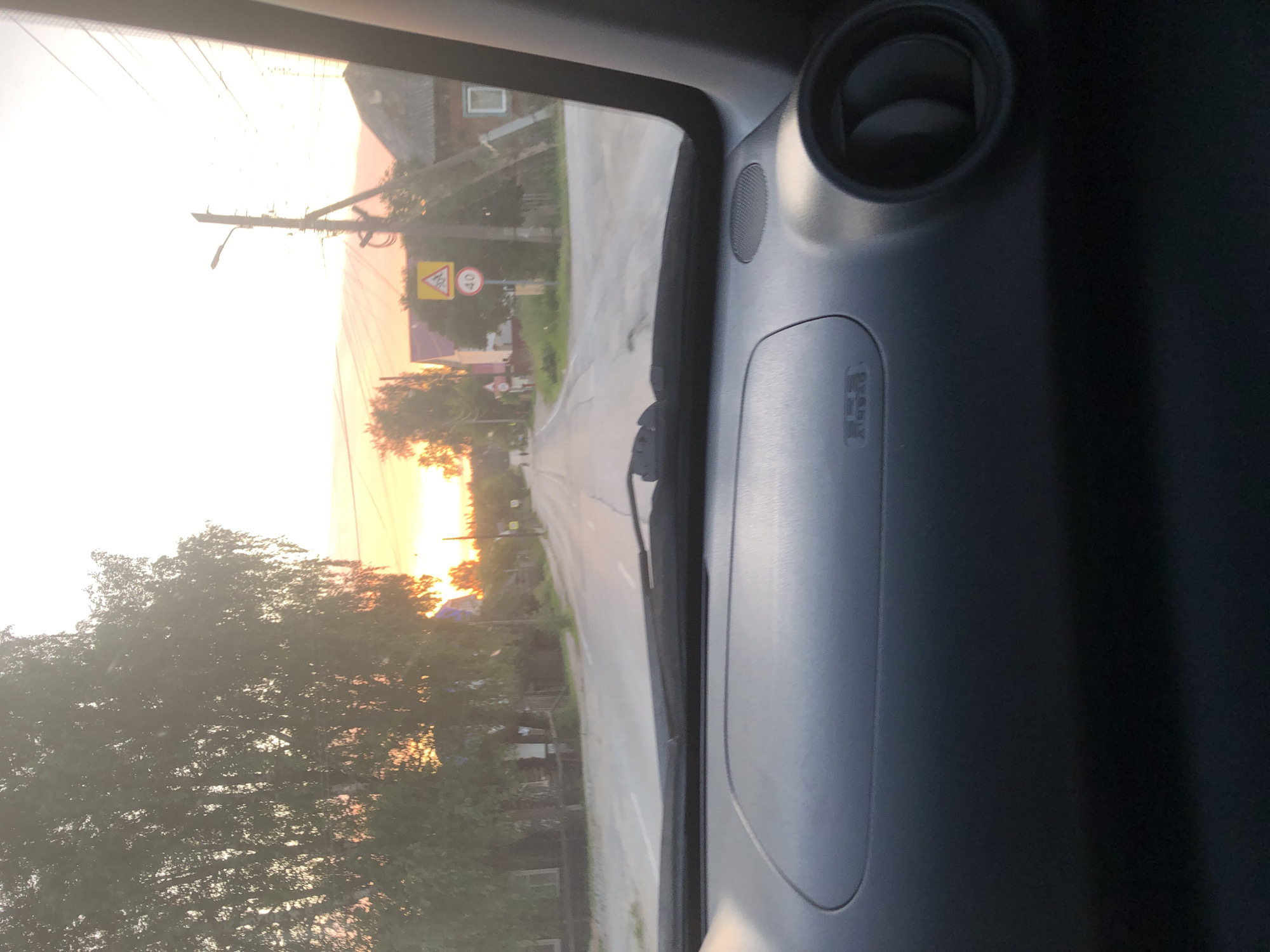 The image shows a view from inside a car, looking out the windshield at a rural road in Solikamsk, Russia. The scene is illuminated by the warm glow of a sunset, casting long shadows and highlighting the golden hues of the sky. On the left side of the road, trees with dense green foliage line the path, their branches interspersed with glimpses of houses and buildings, suggesting a village or small town setting.
A utility pole with wires extends upwards near the side of the road, and a yellow triangular warning sign with a black symbol is visible, along with a circular sign indicating a speed limit of 40. The road itself is paved and curves gently into the distance, with white lines marking the lanes. The interior of the car is visible on the right, featuring a dark gray dashboard with a circular air vent and a panel with "SRS AIRBAG" text printed on it. The overall impression is one of a peaceful drive through the countryside during a beautiful sunset. The time of day is clearly evening, and the weather appears clear.