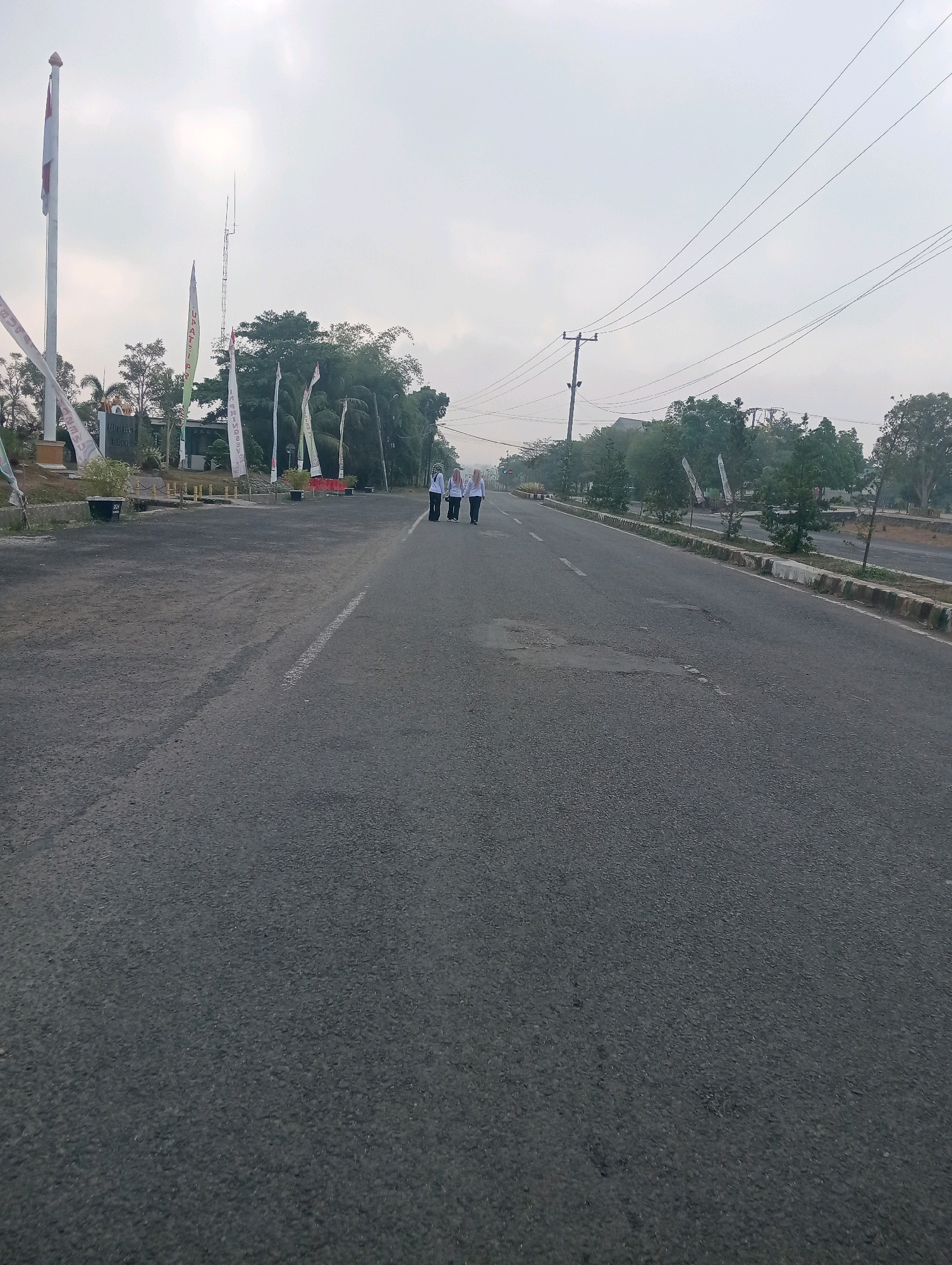 The image depicts a wide, paved road in Sidoharjo, Indonesia, under an overcast sky. Three individuals, dressed in white shirts and dark bottoms, are walking away from the camera on the right side of the road. The road surface is dark gray asphalt with visible white lane markings and some patchy repairs. To the left of the road, there is a paved area with a flagpole flying an Indonesian flag, several other flags, and some foliage. In the background, trees line the right side of the road, and power lines are visible overhead. The overall scene suggests an outdoor, possibly public, area in a semi-rural or suburban environment.