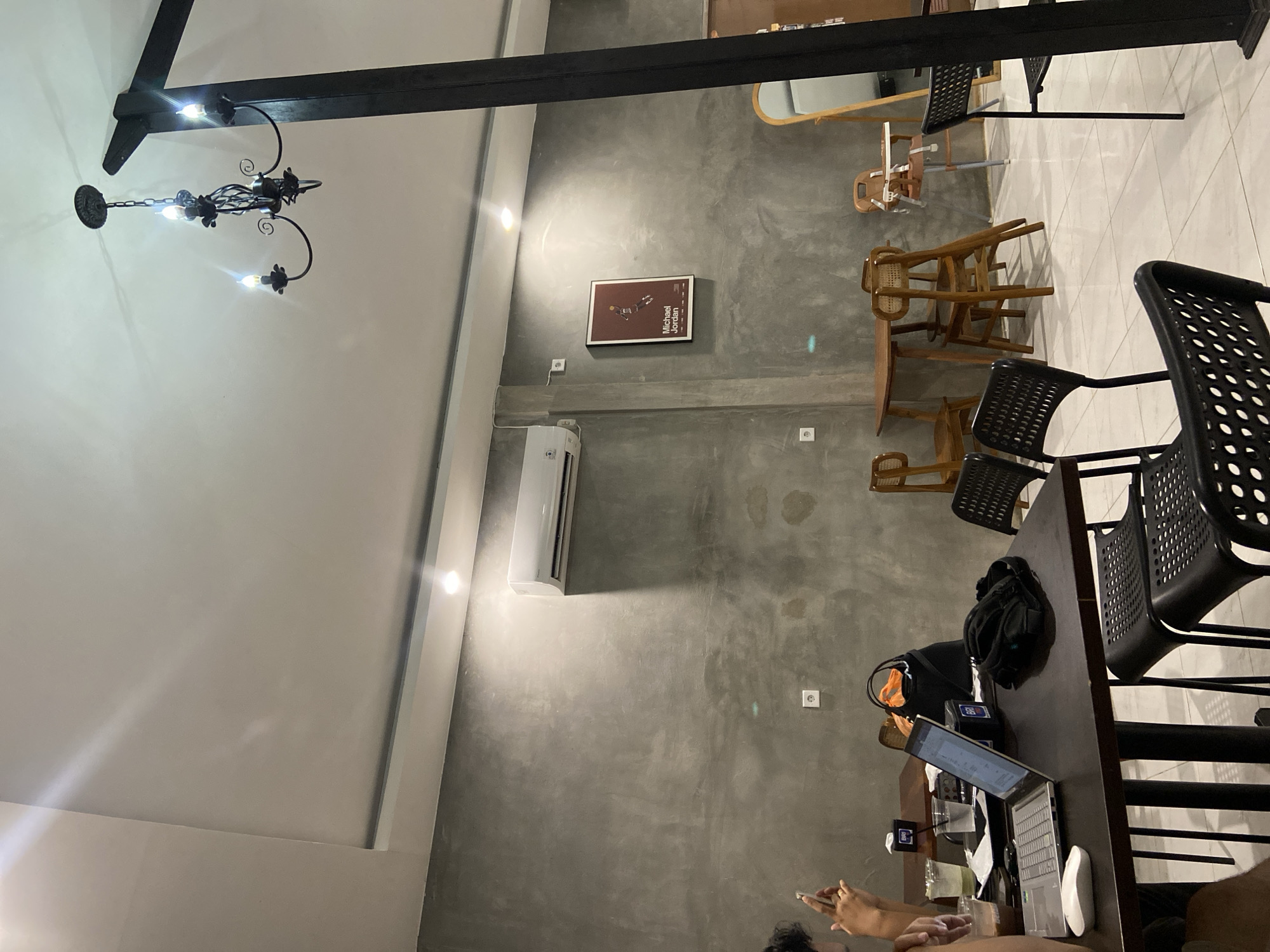 The image shows an indoor scene, likely a cafe or restaurant, with a contemporary, industrial aesthetic. The walls are a rough, textured gray concrete, and the ceiling is white. A dark wooden beam spans across the upper part of the image, from which hangs a decorative black chandelier.
The space is furnished with several wooden chairs, some with woven seats, and black perforated chairs. A long, dark wooden table is set up, with a laptop open, a computer mouse, and drinks on it. A backpack and an orange bag are also on the table. Two people are partially visible, one appearing to use a phone and another working on the laptop.
On the gray wall, a framed poster with the text "Michael Jordan" is visible. An air conditioning unit is mounted on the wall, suggesting a climate-controlled environment. The lighting appears to be artificial, with the chandelier and perhaps spotlights casting light. The overall atmosphere suggests a relaxed, perhaps studious or work-oriented environment. The location context indicates Sekar Mawar, Indonesia, but there are no specific visual cues to confirm this beyond the provided information. The time of day is not discernible.