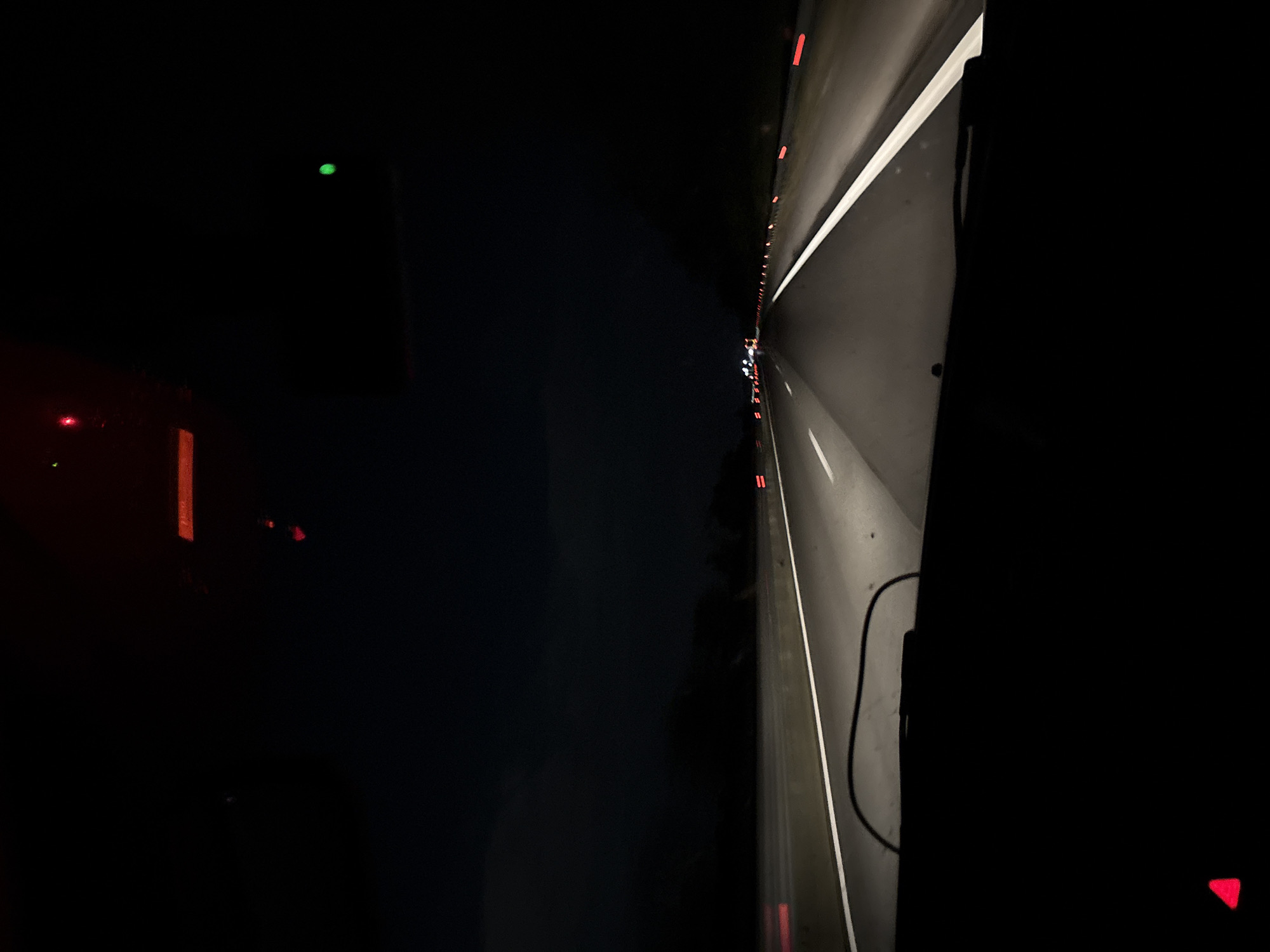 The image depicts a nighttime view from a moving vehicle, possibly a car or truck, on a highway. The right side of the image shows the highway illuminated by headlights, including white lane markings and possibly other vehicles' tail lights in the distance. The left side of the image is dark, with glimpses of what appears to be a dashboard or interior of the vehicle, including faint lights. The scene suggests nighttime travel on a well-lit highway, but lacks specific identifying details to determine a location in Manole, Bulgaria.