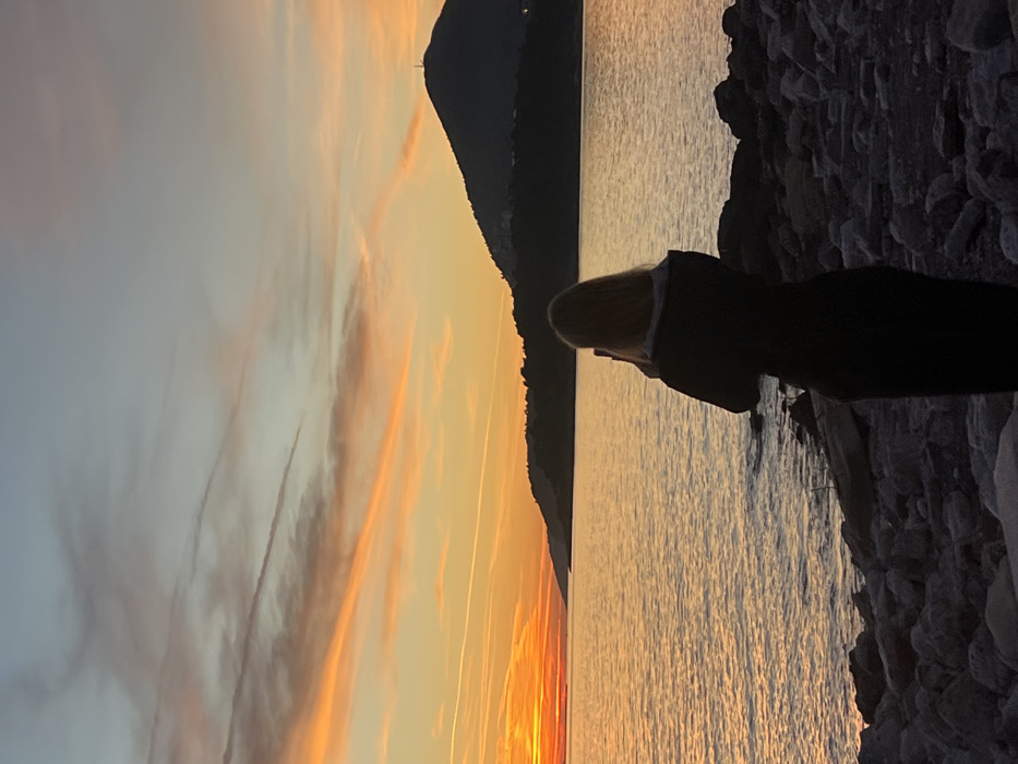 A scenic landscape shows a person standing at the edge of a rocky shore, gazing out at a body of water reflecting a vibrant sunset. Silhouetted mountains rise in the background. The sky is painted with hues of orange and yellow, streaked with clouds. The water is calm, mirroring the sky's colorful display. The figure appears contemplative, enjoying the peaceful moment.