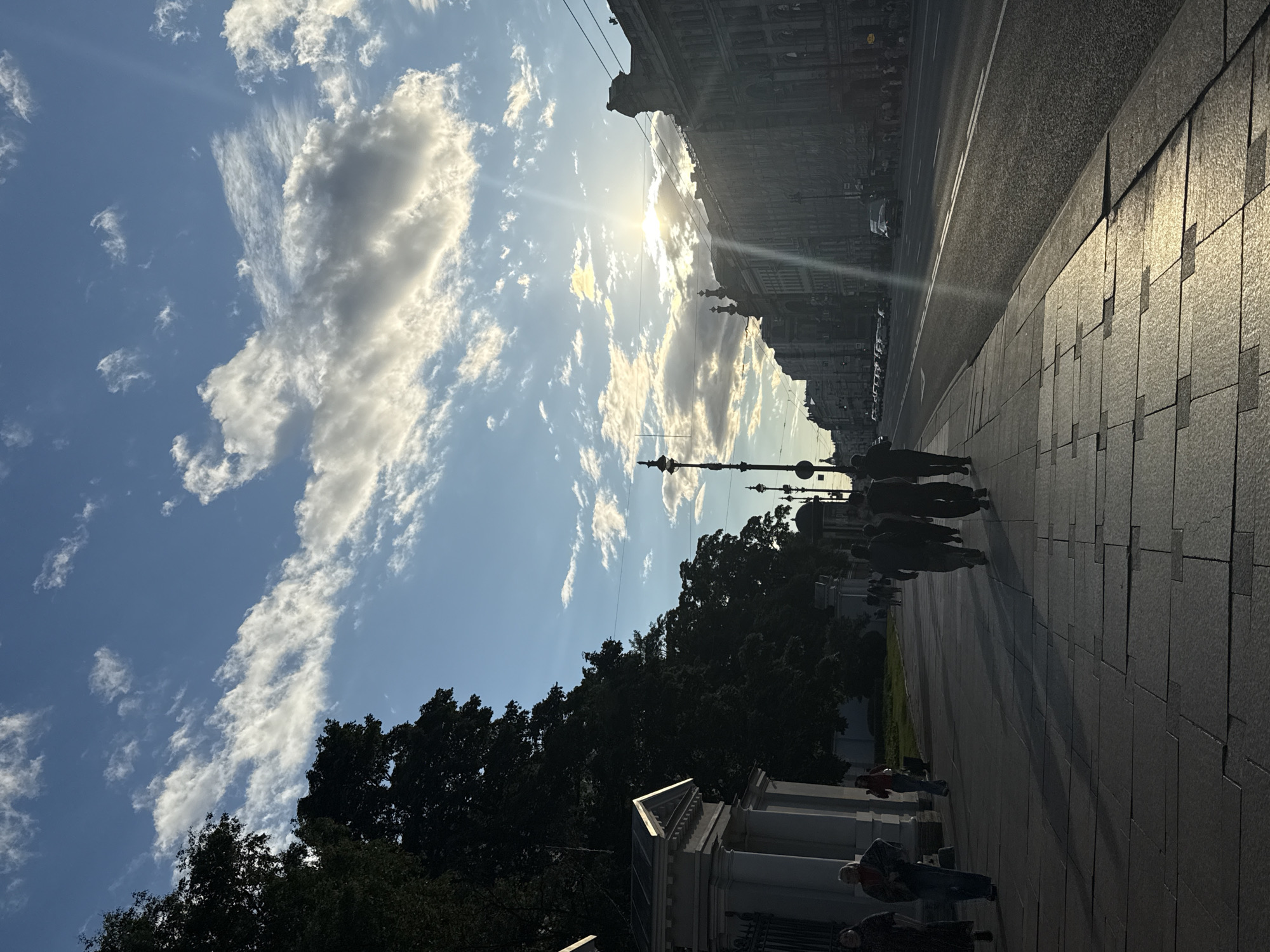 The image depicts a street scene in Saint Petersburg, Russia, likely during a late afternoon or early evening due to the low angle of the sun and the long shadows. On the left side of the frame, a paved sidewalk runs alongside a street. Several people are walking along the sidewalk, some in silhouette. To the left of the sidewalk, there is a white neoclassical building with columns and ornate details, partially obscured by large, dark green trees. Across the street, on the right side of the frame, are imposing, dark-colored buildings with classical architectural features, typical of Saint Petersburg's historic center. The street itself appears to be a multi-lane road with parked cars visible in the distance. The sky above is blue with scattered white clouds, and the setting sun is visible, casting a warm glow and a prominent sunbeam across the scene, reflecting off the wet or polished pavement of the sidewalk.