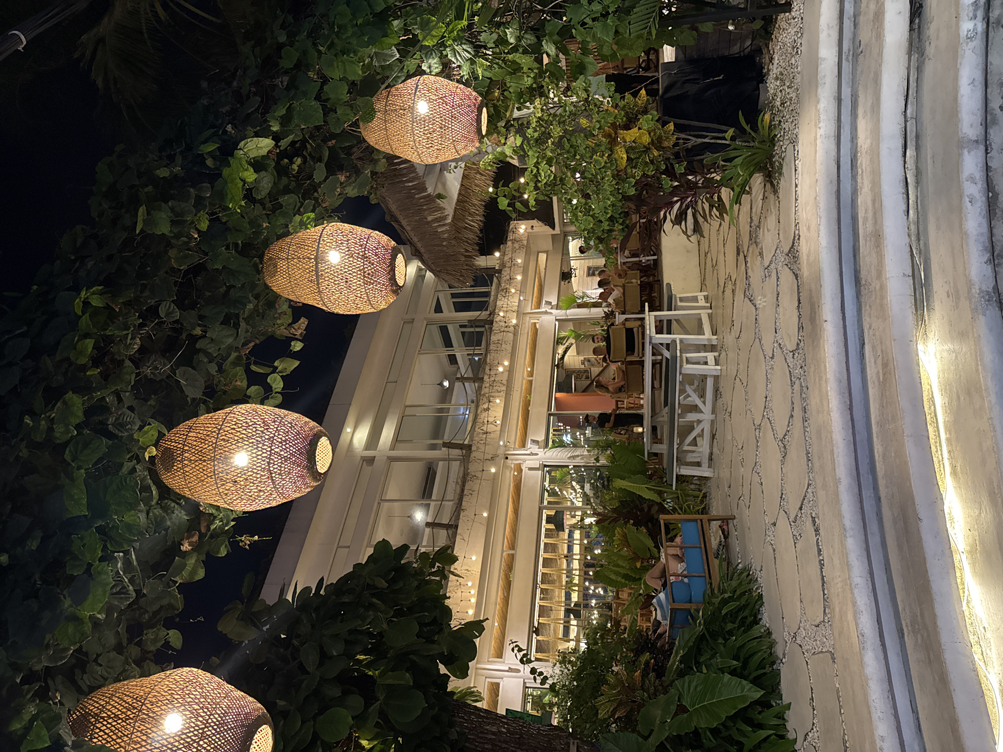 The image depicts an outdoor dining area at night, likely in Kuta, Indonesia, given the tropical vegetation and the architectural style. Hanging from trees and vines are several woven pendant lights, casting a warm glow. In the foreground, there are concrete steps illuminated from below. The middle ground features a patio paved with irregular stone slabs interspersed with gravel. To the left, a person reclines on a blue-cushioned bench amidst lush green foliage. Further back, a white picnic table with benches is visible. Beyond that, a two-story building with large windows and an open-air ground floor serves as a restaurant. String lights are draped along the building's exterior. Inside and outside the restaurant, patrons are seated at tables, engaged in dining and conversation. The overall ambiance is relaxed and inviting, characteristic of a tropical evening.