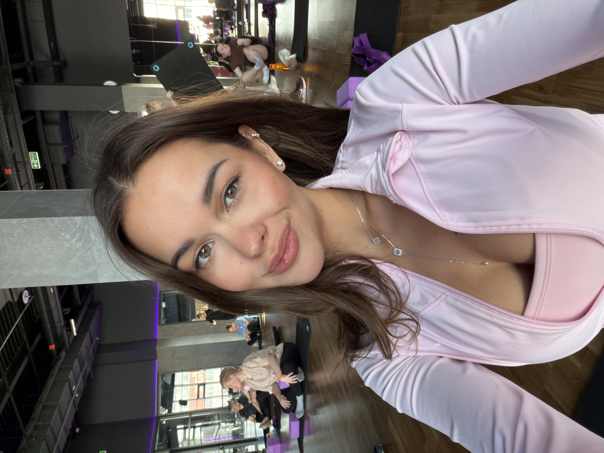 Here is a description of the image:
The image shows a woman taking a selfie in what appears to be a yoga studio or fitness center. The woman has brown hair and is wearing a light pink athletic top. She has a necklace on. In the background, there are other people doing yoga or stretching on mats. The gym or studio seems to have a modern design with large windows and concrete pillars. The location is in Krasnoyarsk, Russia.