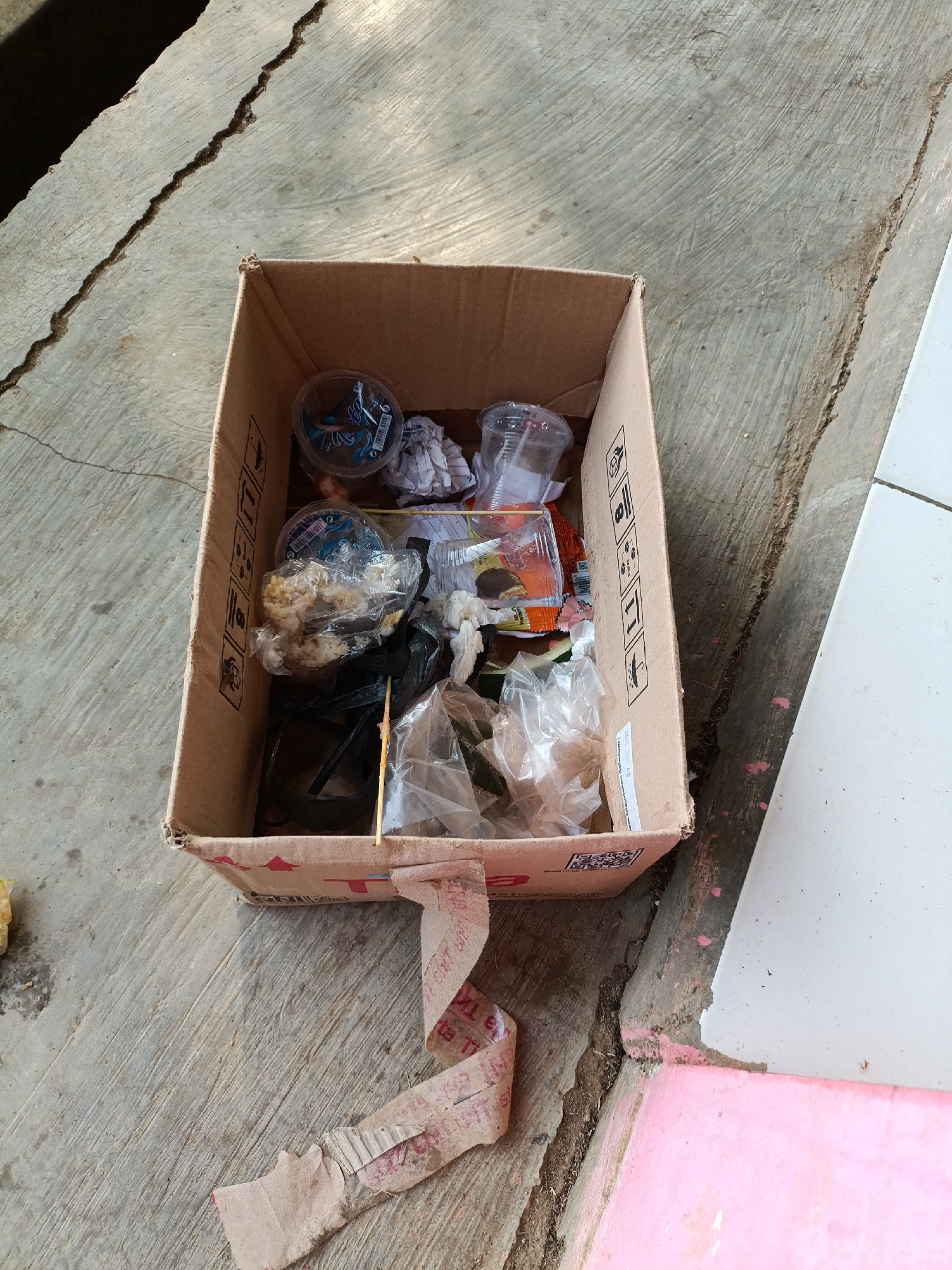 A cardboard box filled with miscellaneous trash, including plastic cups, wrappers, and what appears to be food scraps, is placed on a cracked concrete surface. The trash is varied in color and texture. The box itself is slightly worn. The surrounding environment suggests an outdoor setting, possibly in an alleyway or near a building, with a pink-painted surface visible in the lower right corner. The scene appears to be located in Sumberejo, Indonesia.