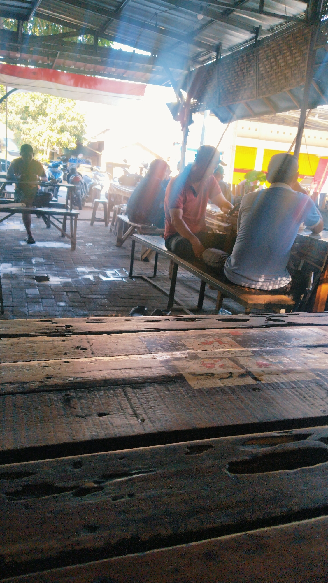 A group of men are seated at tables in an open-air eatery located in the city of Besuki, Indonesia. The eatery appears to have a metal roof supported by visible beams, and a red and white Indonesian flag hangs prominently from the ceiling. Sunlight streams through the structure, creating lens flares and illuminating dust particles in the air.
In the foreground, a rustic wooden table dominates the view, its worn surface textured with visible grain and small holes. Behind this table, several individuals are seated on benches at other tables. One man in a green t-shirt is visible in the background to the left, seated at a table. In the mid-ground, a man in a red polo shirt is seated, looking down towards a table. To his right, another man in a light blue t-shirt is also seated. In the distance, more tables and individuals are partially visible, along with motorcycles parked to the left. The ground is paved with brick, and some areas appear wet, reflecting the light. The overall impression is of a casual outdoor dining establishment.