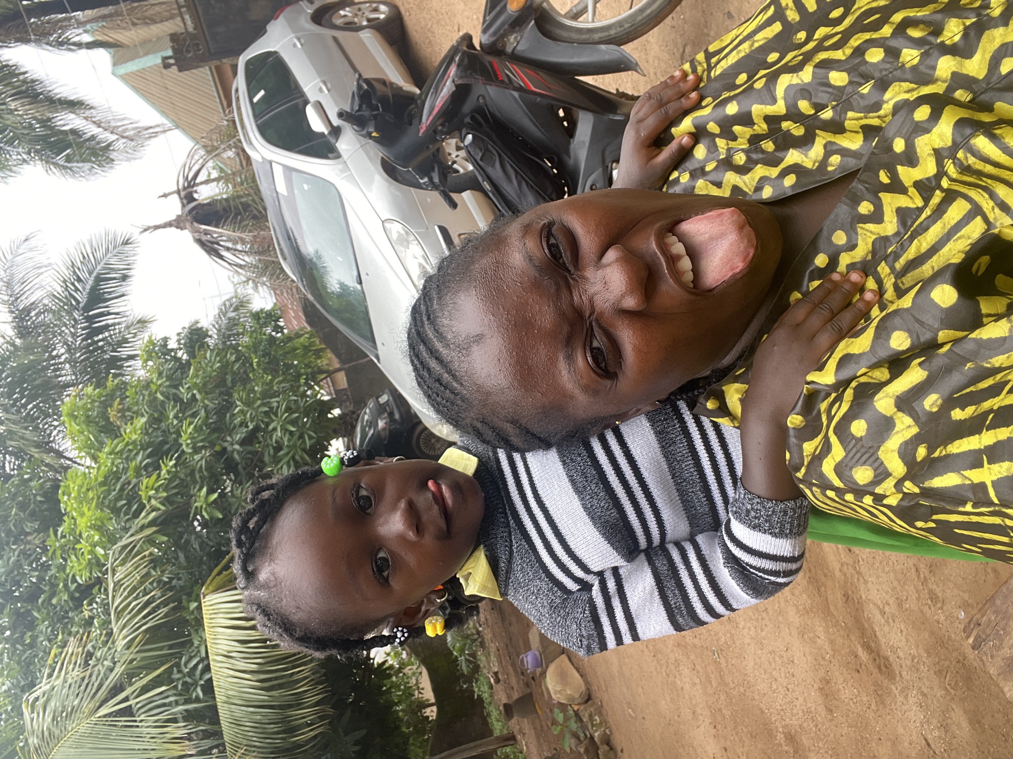 The image captures a close-up selfie taken outdoors in a rural or semi-rural setting in Akwanga, Nigeria. In the foreground, a woman is making a face, sticking her tongue out at the camera. She is wearing a yellow and black patterned garment. Behind her, a young girl is peeking over her shoulder, also sticking her tongue out. The girl is wearing a black and white striped sweater and a green skirt, with colorful beads in her braided hair. In the background, a silver car and a black motorcycle are visible, along with lush green foliage and some buildings, suggesting a residential area. The ground is covered in dirt.