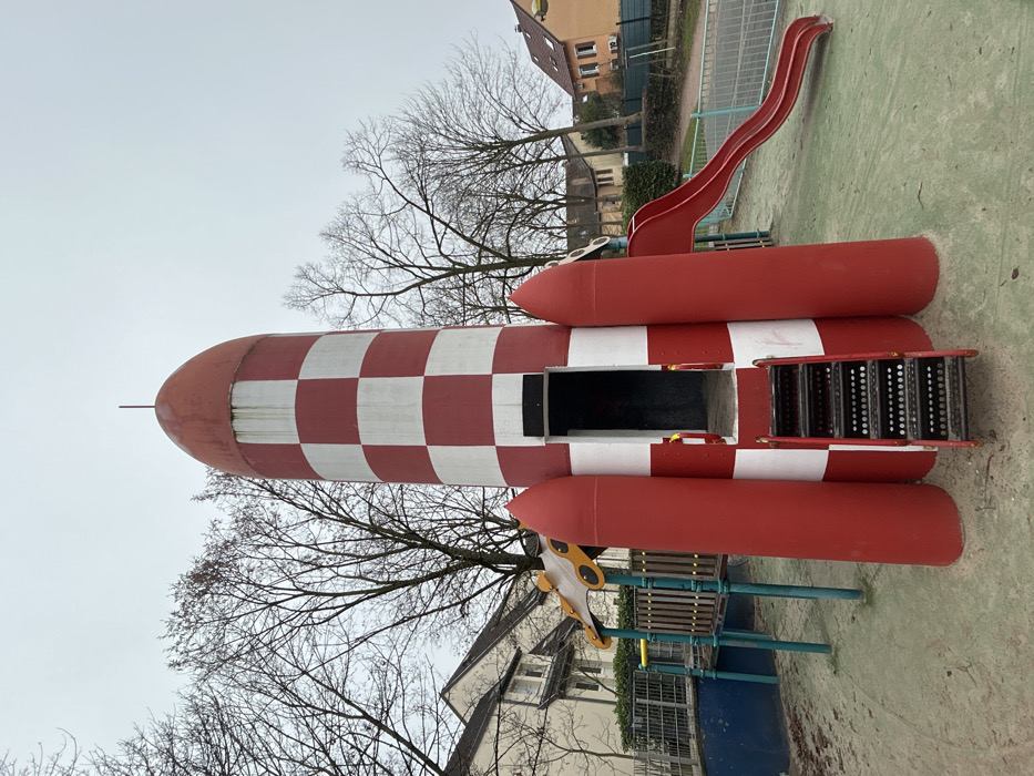 An outdoor playground in Schiltigheim, France, features a prominent rocket-shaped play structure under an overcast sky. The central object is a tall, cylindrical rocket, predominantly red and white, with a checkered pattern on its main body and a solid red conical top. Three red booster-like fins are attached to its base. A black metal staircase with red handrails leads into an open, dark entrance at the rocket's front. To the right of the rocket, a red plastic slide curves down to the ground. To the left, a portion of an adjacent playground unit, featuring a wooden platform, yellow canopy, and blue support posts, is visible next to a blue-painted barrier. The ground consists of a light green, possibly soiled, soft fall surface. In the background, numerous bare deciduous trees stand between residential buildings with light-colored and orange facades. A metal fence separates the playground area from the adjacent properties. No individuals are present in the scene.