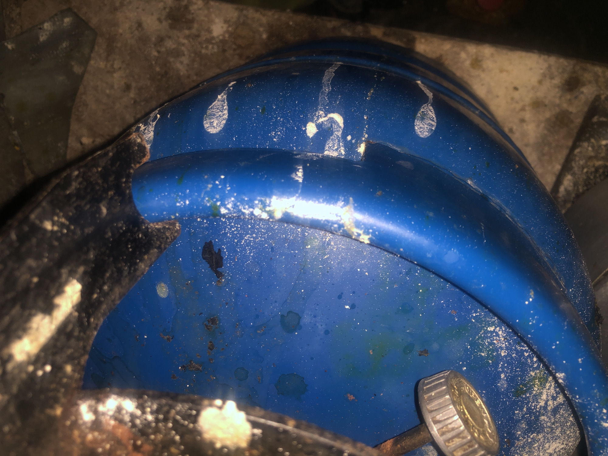 The image depicts a close-up view of a blue, curved object, possibly a container or part of a machine, showing signs of wear and tear. The surface is marred by white splatters, some appearing like dried liquid, and there are areas of rust and what looks like dirt or debris. A metallic knob with ridged edges is visible on the left side. The background is out of focus, suggesting a rough, possibly concrete or dirt surface. The lighting is dim, casting shadows and highlighting the textures of the object. The image does not contain any discernible features that would allow for the identification of its location within Jalingo, Nigeria, nor does it show any people, actions, or interactions.