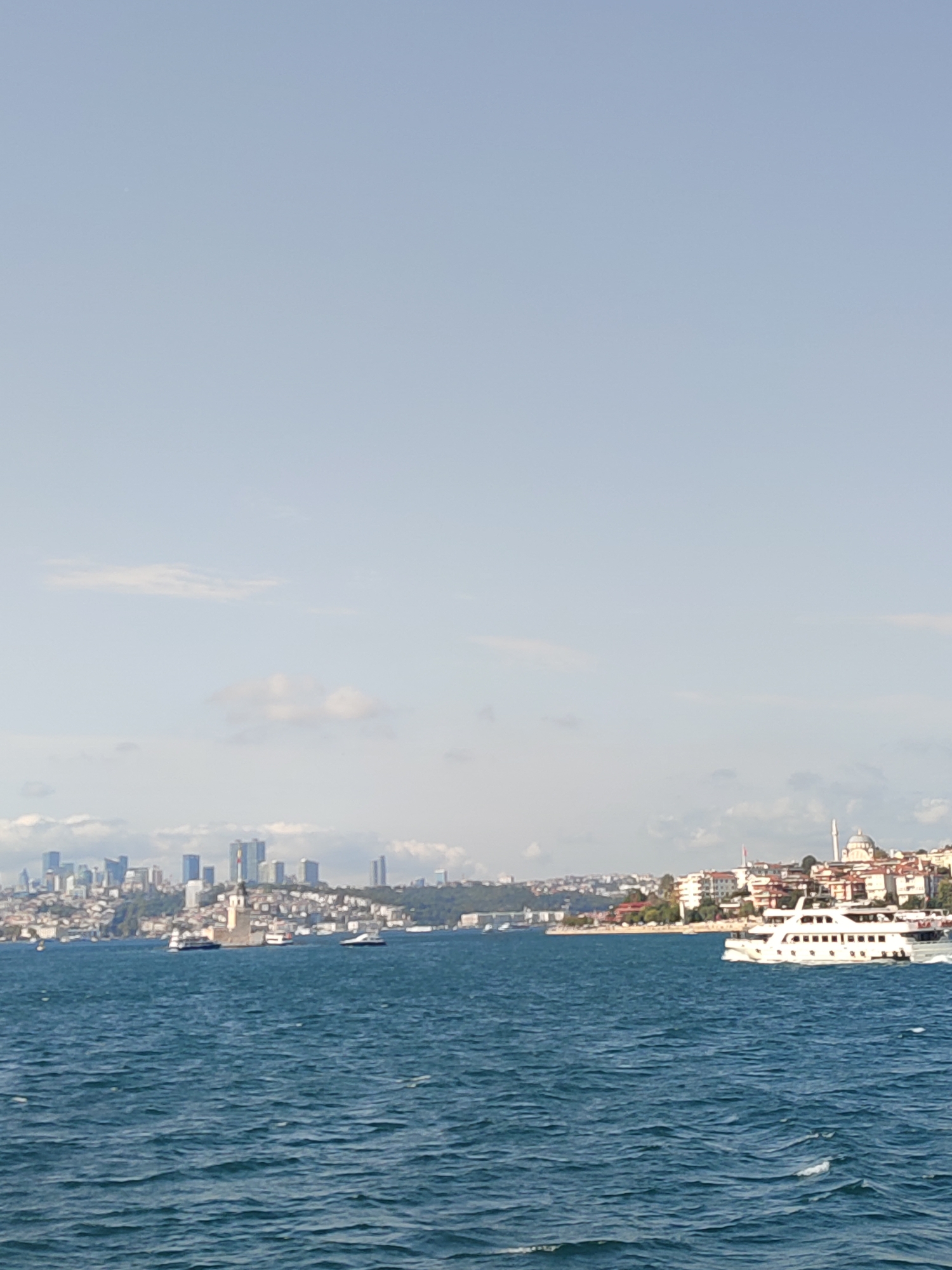A wide shot captures the Üsküdar coastline in Istanbul, Turkey, with the blue Bosphorus strait dominating the foreground. The water is choppy, with small whitecaps visible. On the left side of the image, a cluster of modern skyscrapers forms the city skyline. In the middle ground, the Maiden's Tower, a historical landmark, stands on a small island. Several boats, including a white passenger ferry, are navigating the strait. The background depicts a densely populated urban area with numerous buildings and the distinctive dome and minaret of a mosque. The sky is a pale blue with scattered clouds.