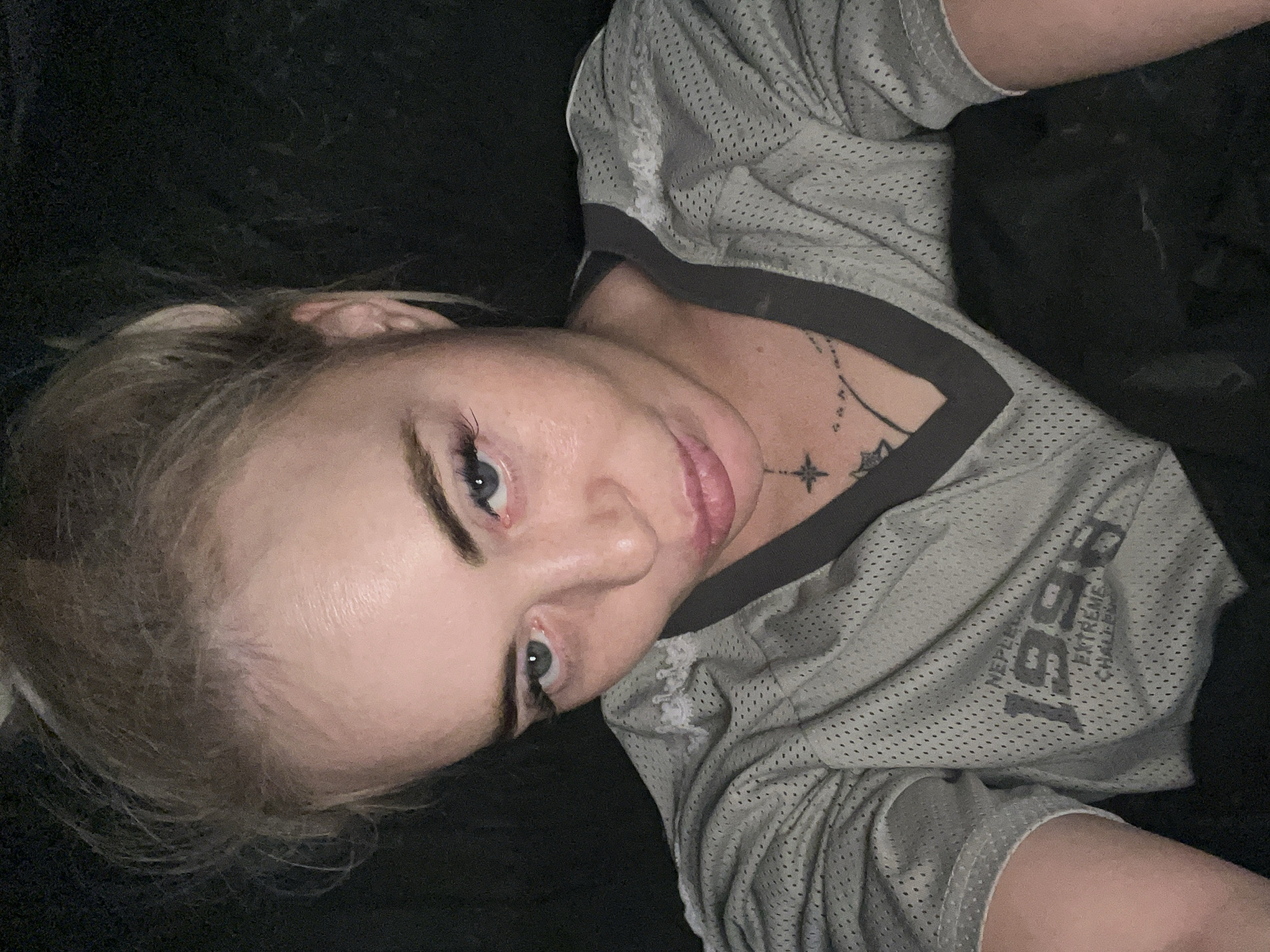 A close-up, over-the-shoulder selfie shows a fair-skinned woman with blonde hair pulled up in a bun. She is wearing a grey, mesh athletic jersey with the number "1996" and the words "EXTREME CHALLENGE" visible on the front. A dark, star-shaped tattoo is visible on her chest, partially covered by the jersey's neckline. The background is dark and indistinct. The woman's expression is serious, with her eyes looking directly at the camera.