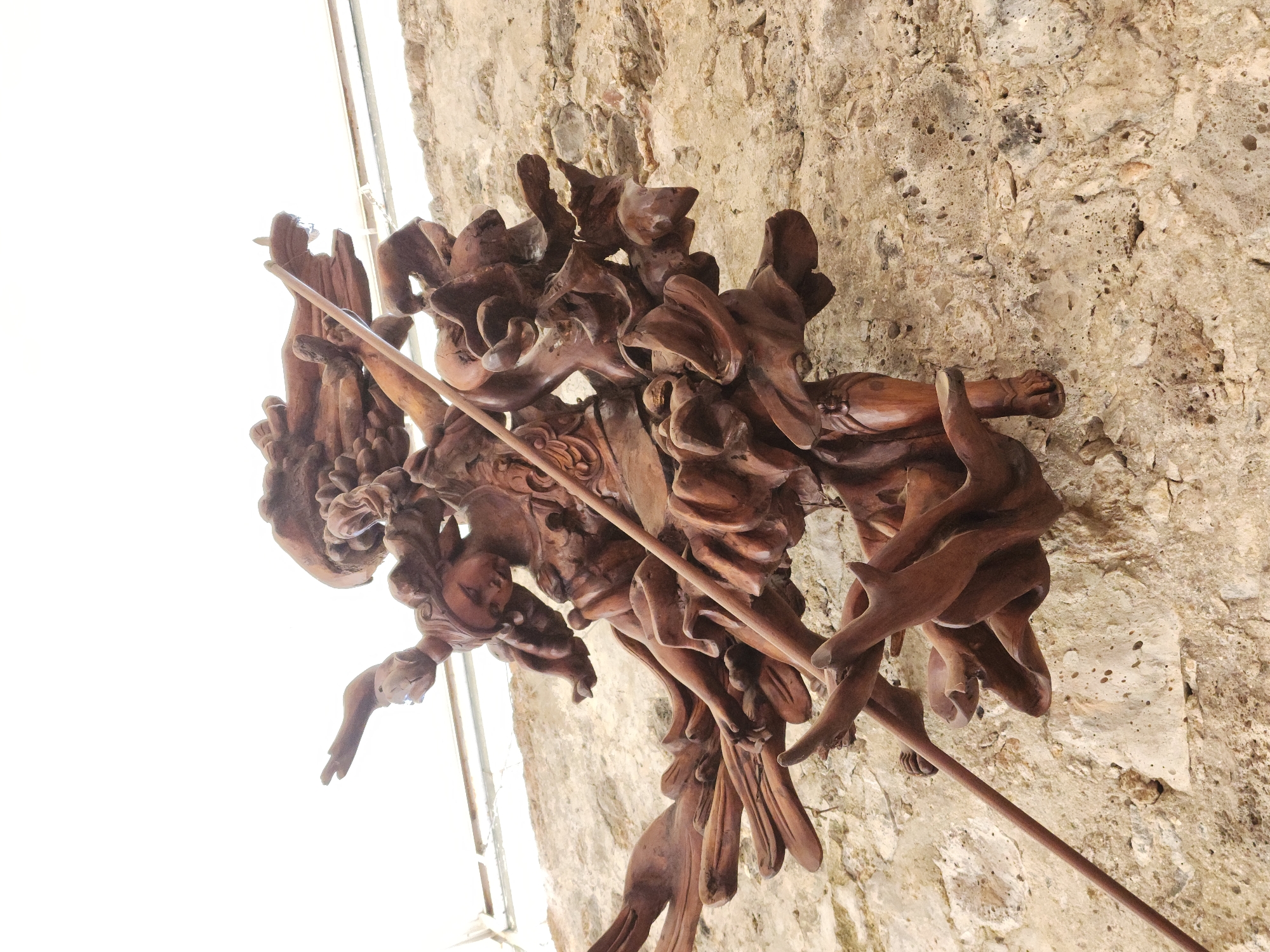 The image depicts a wooden sculpture, possibly depicting figures or forms, mounted on a stone wall. The sculpture is brown and appears intricately carved. The wall is light-colored and textured. The location is Unknown, Unknown.