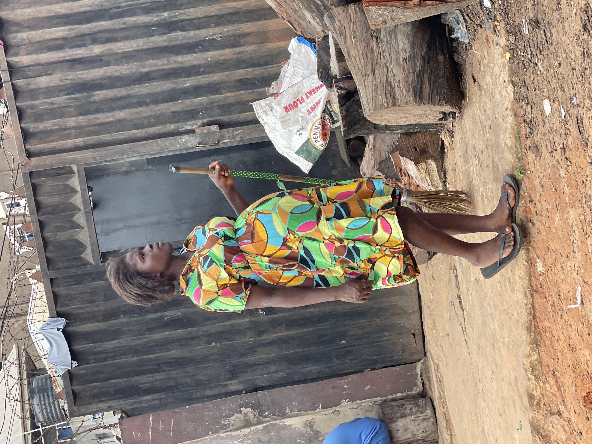 A woman is sweeping a dirt area in Nkpor, Nigeria. She is wearing a colorful dress and black flip-flops. The scene is situated outside a corrugated metal structure. Debris and a bag of flour are visible near the structure, and barbed wire is along a fence in the background.